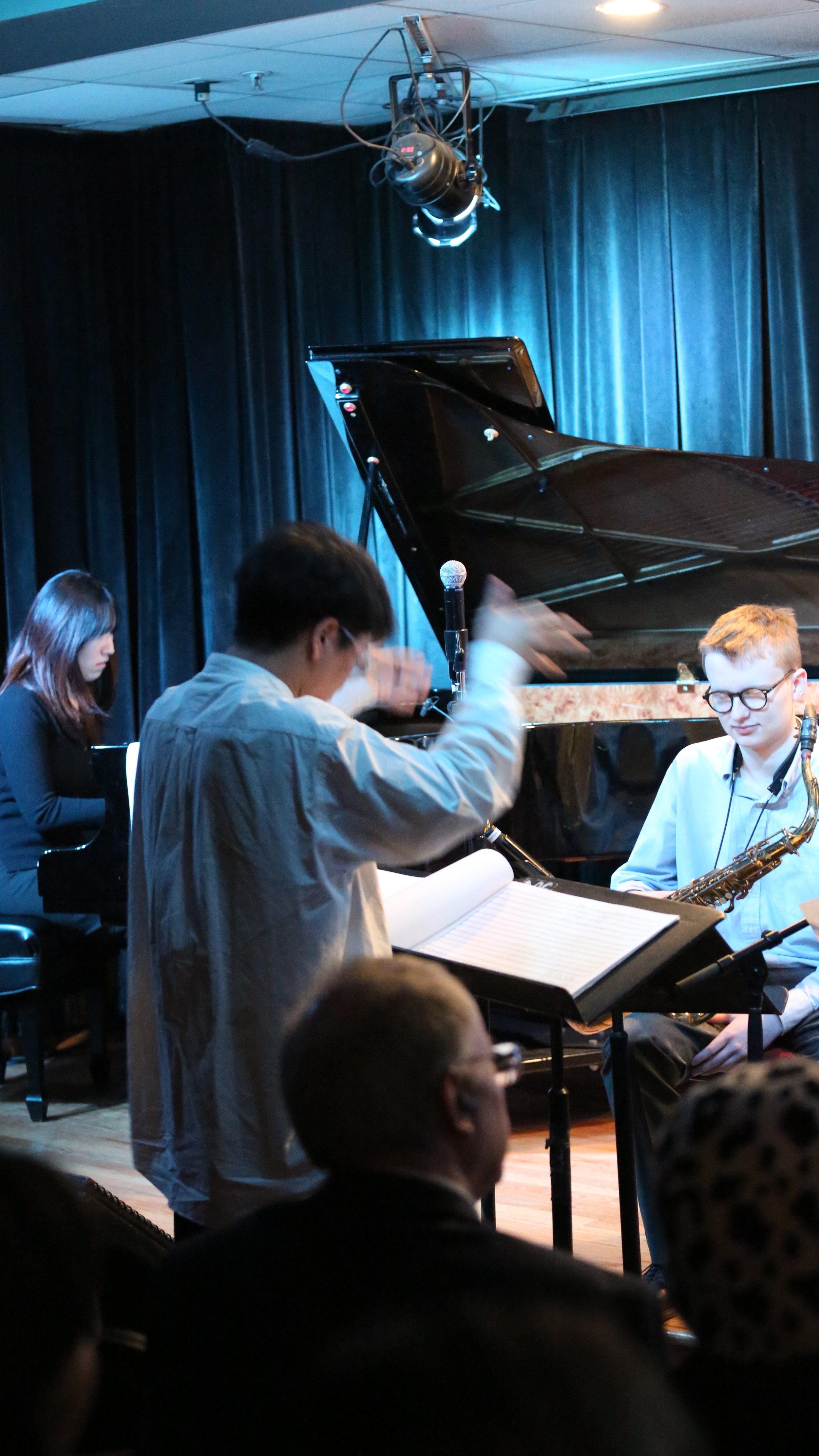 Jazz band performing on stage with musicians playing piano and saxophone, conductor leading, audience watching in a dimly lit venue.