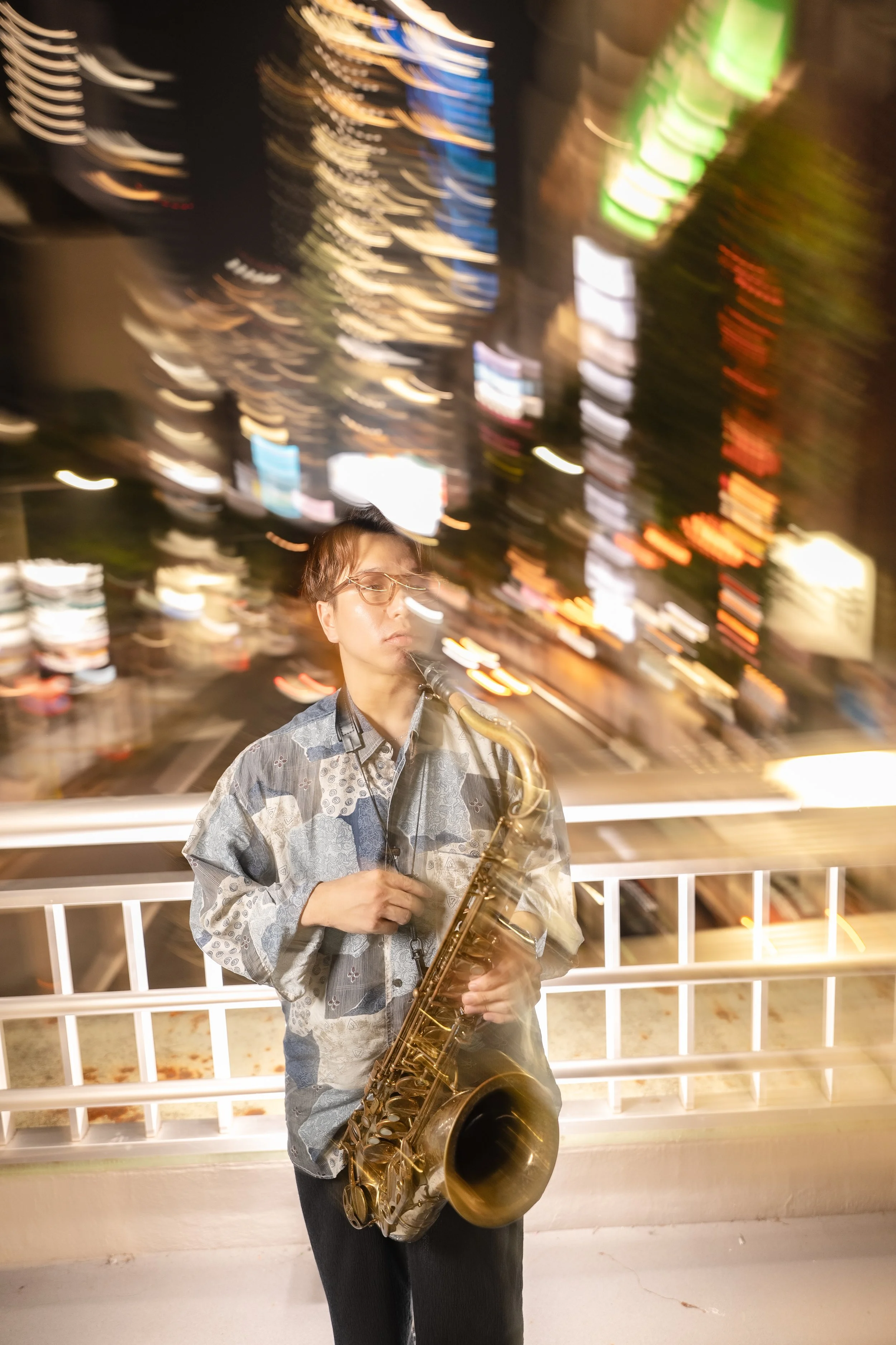 A person with short hair and glasses playing a saxophone at night with city lights blurred in the background.