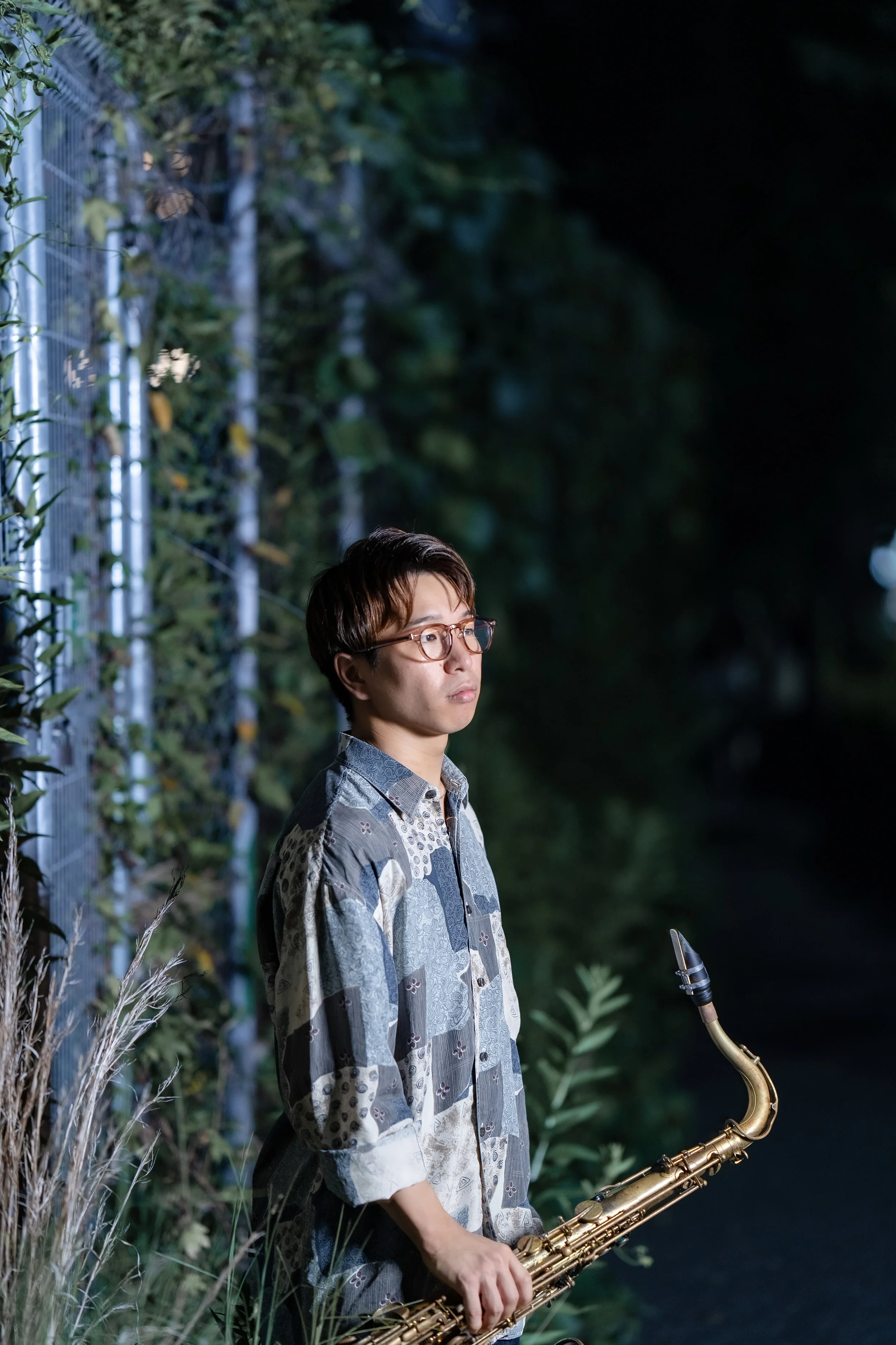 A person with short dark hair and glasses standing outdoors at night, holding a saxophone, with greenery and a metal fence in the background.
