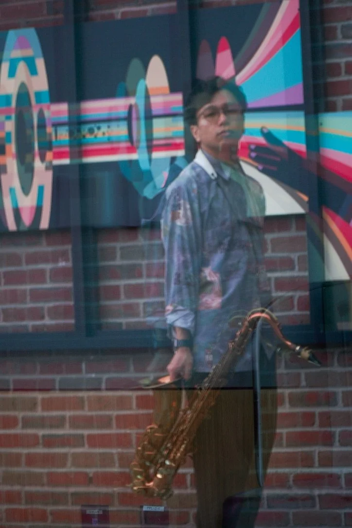 Reflection of a young man holding a saxophone, taking a selfie in a glass window with colorful abstract artwork on the wall behind him.