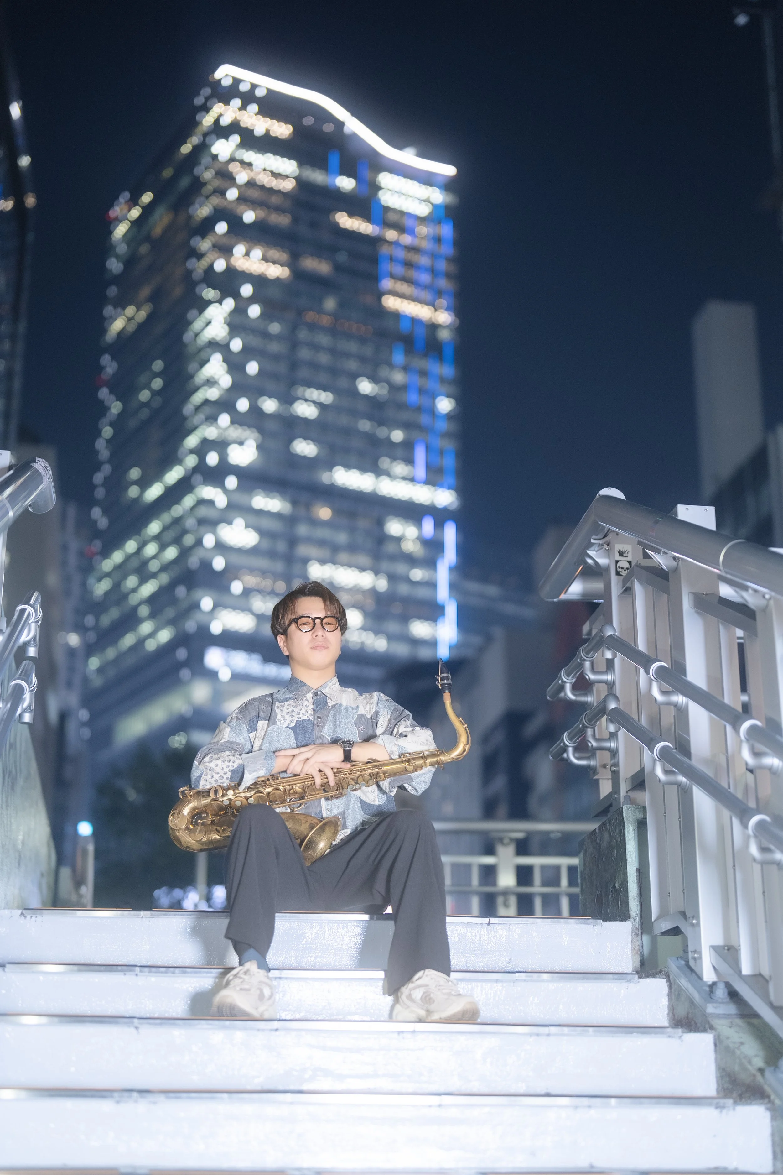 A person sitting on stairs at night with a saxophone, city buildings with illuminated windows and blue lighting in the background.
