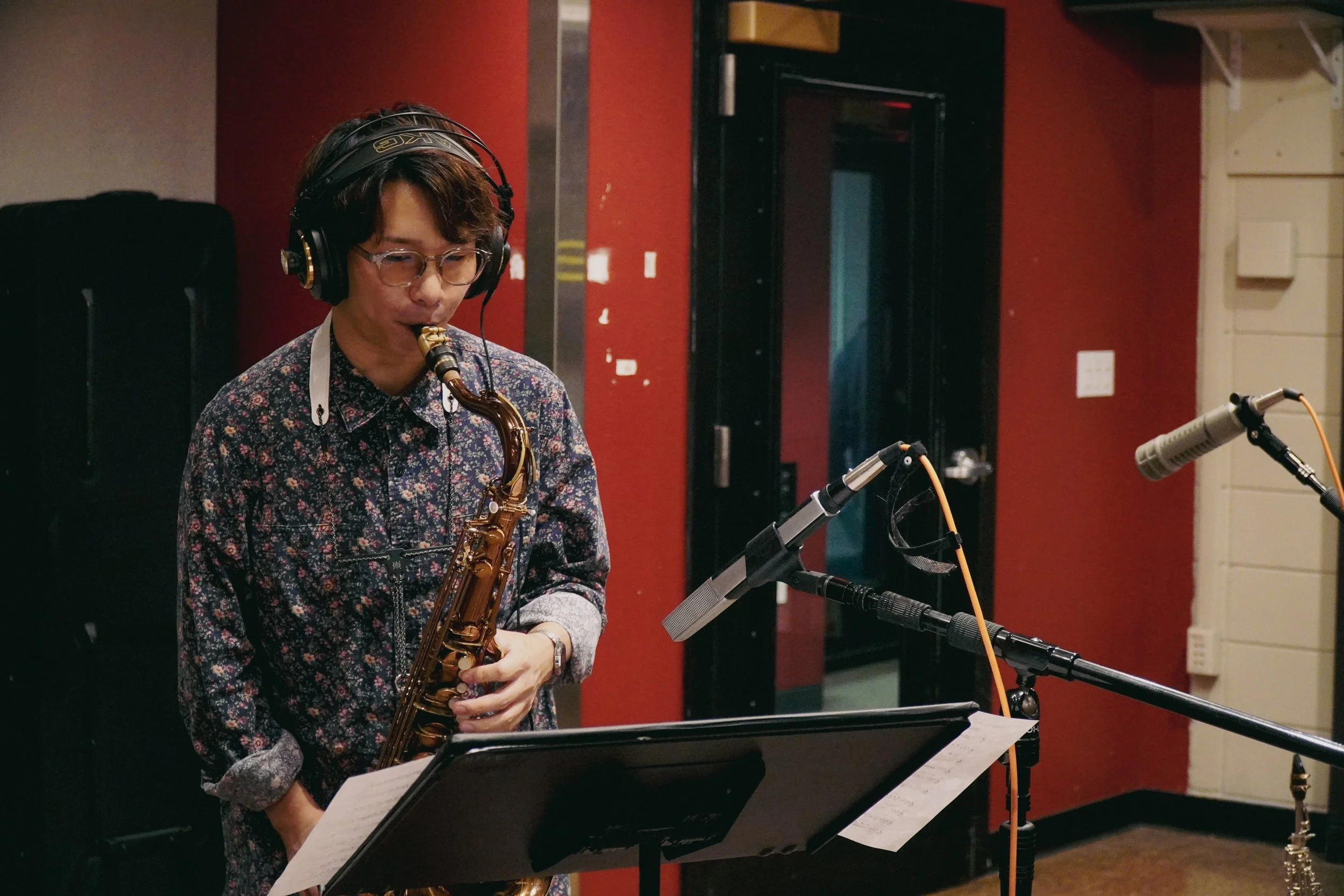 A person with glasses and wearing a floral shirt playing a saxophone in a recording studio.