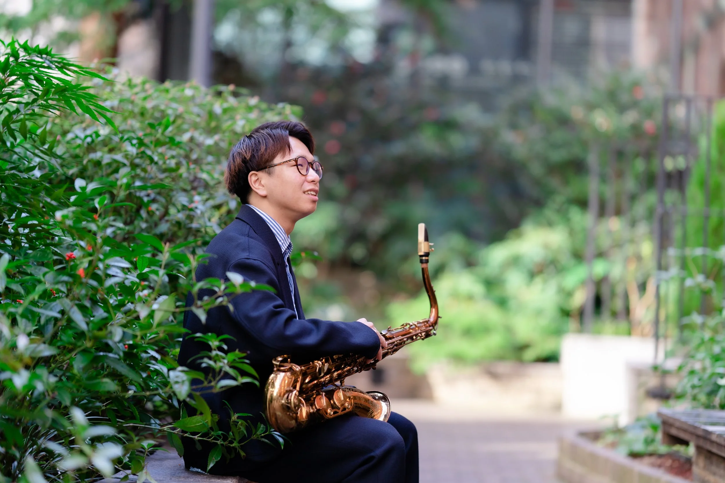 A person wearing glasses, in a dark suit, sitting on a park bench surrounded by greenery, playing a saxophone.