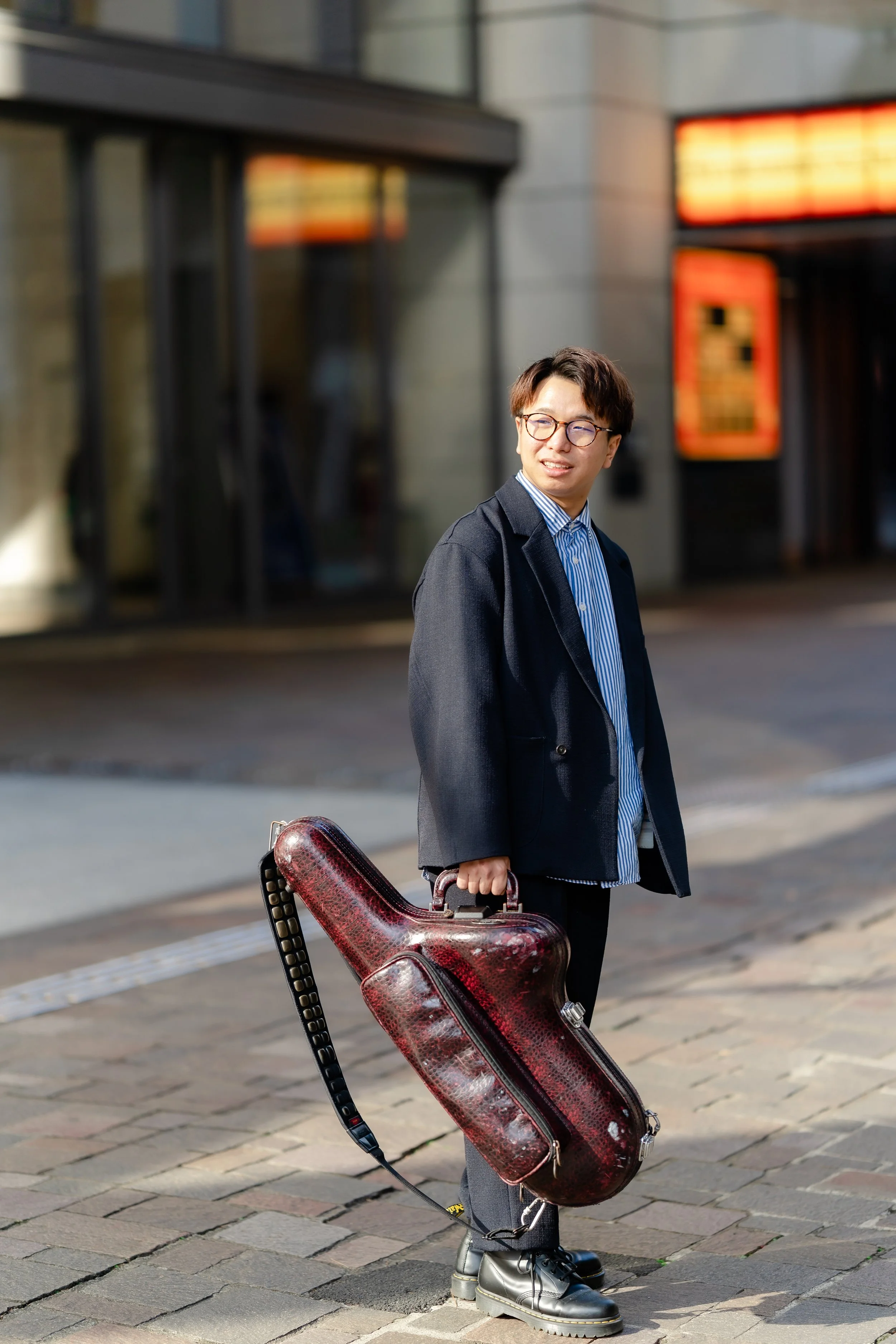 A young man with glasses wearing a grey blazer and striped shirt, standing on a city street with a guitar case in hand, smiling and looking to the side.