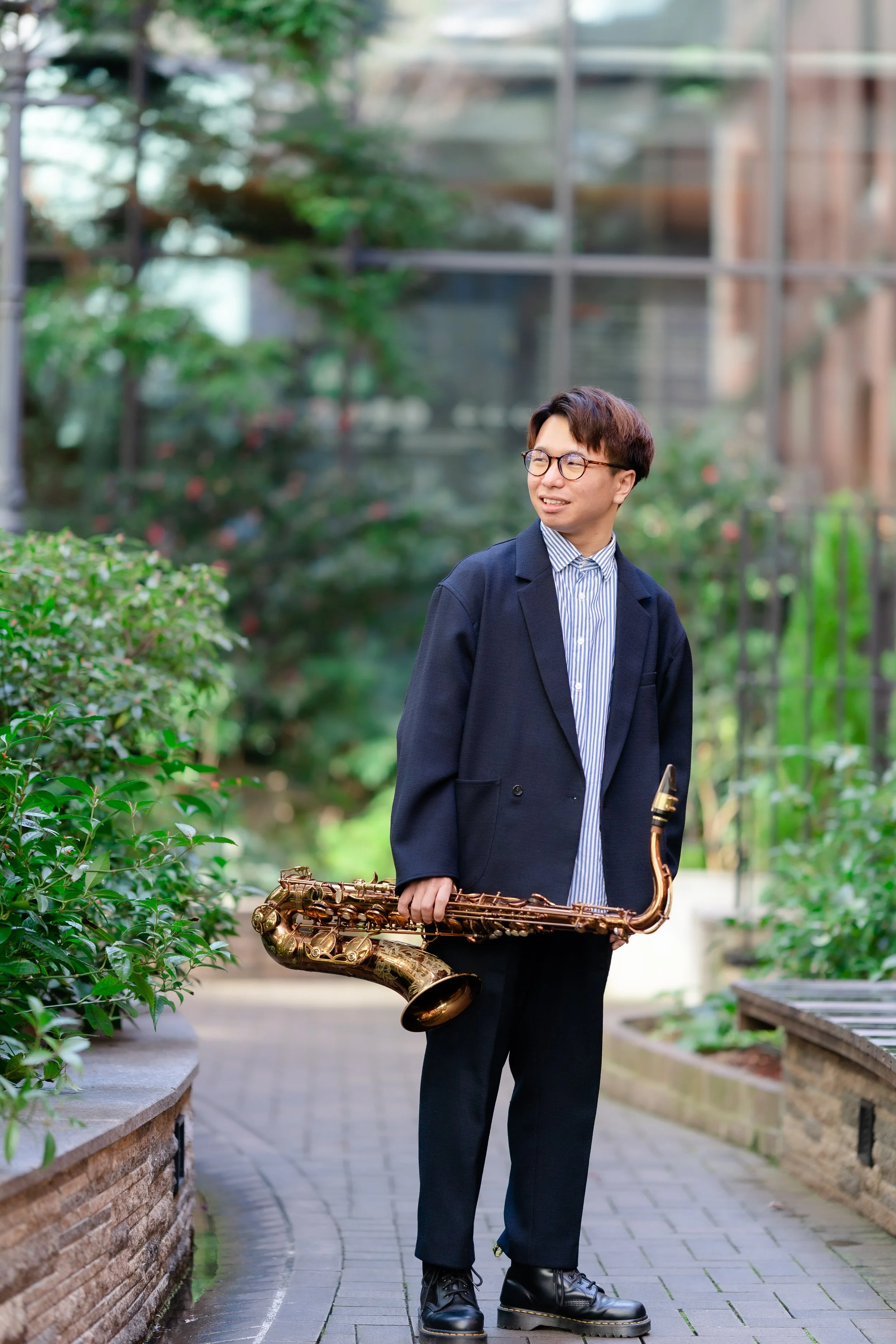 Man in glasses and a dark blazer holding a saxophone, walking outdoors on a paved pathway surrounded by greenery and brick planters.