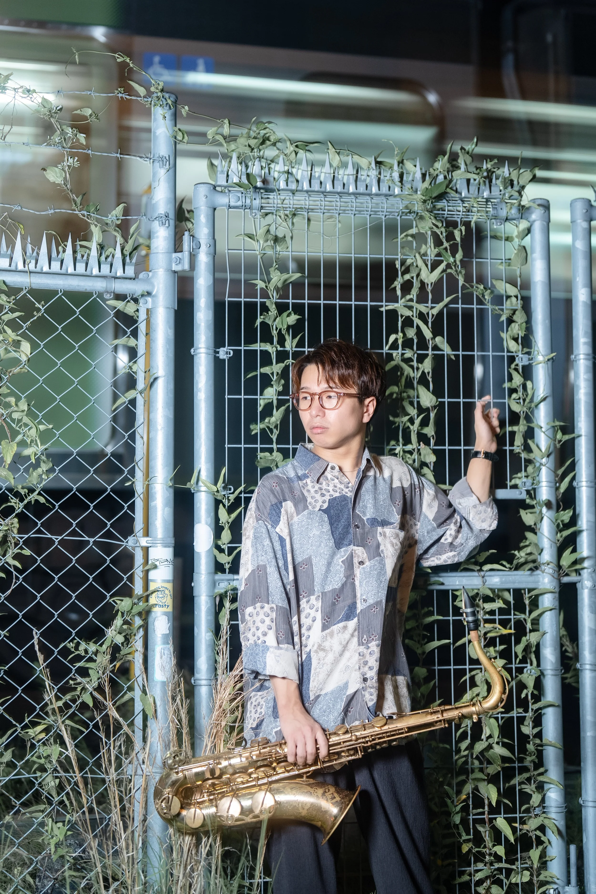 A man with glasses dressed in a patterned shirt and dark pants holding a saxophone, standing next to a chain-link fence with plants growing on it at night.