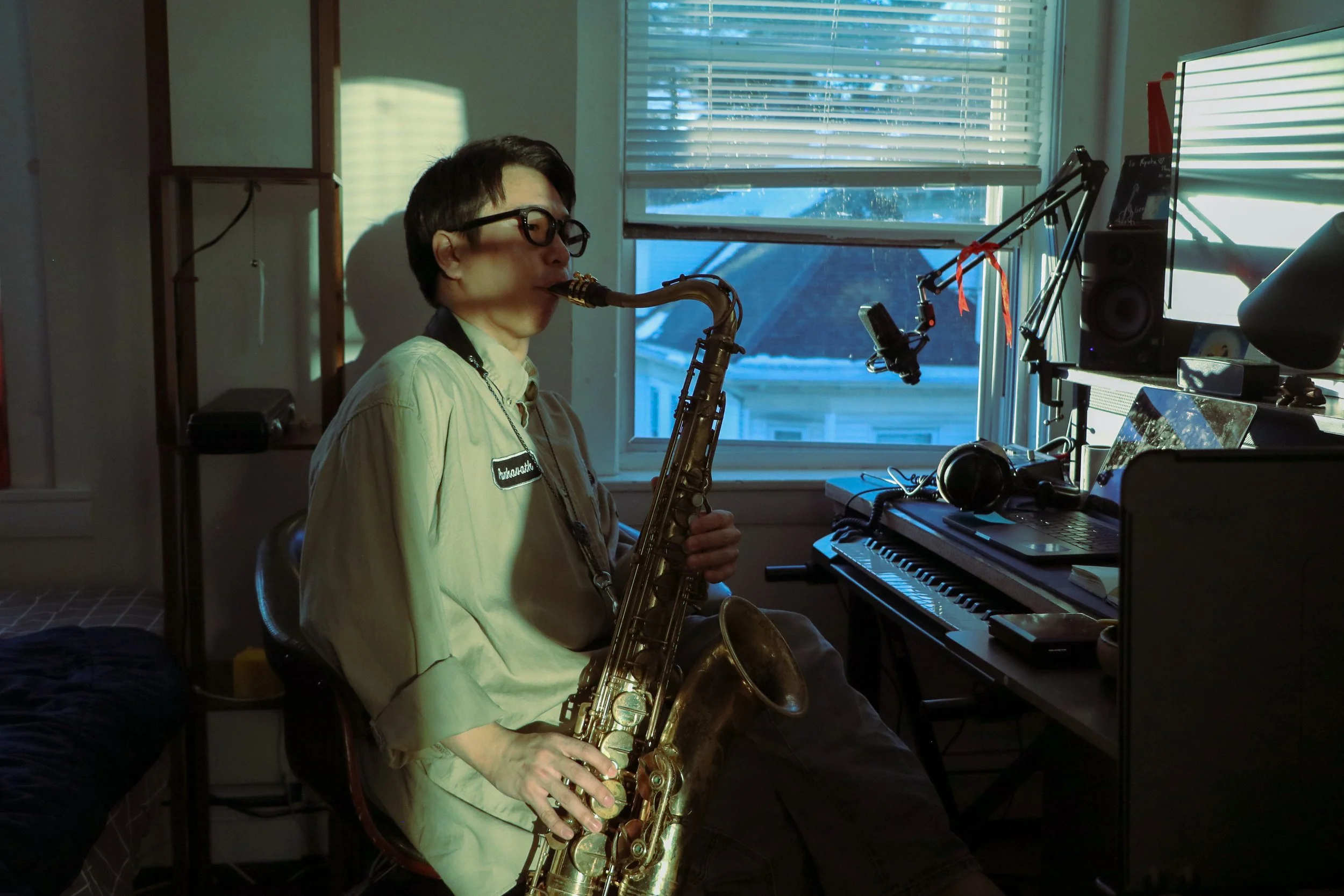 Person with short dark hair and glasses playing a saxophone in a room with a computer setup, window with blinds, and various electronic equipment.