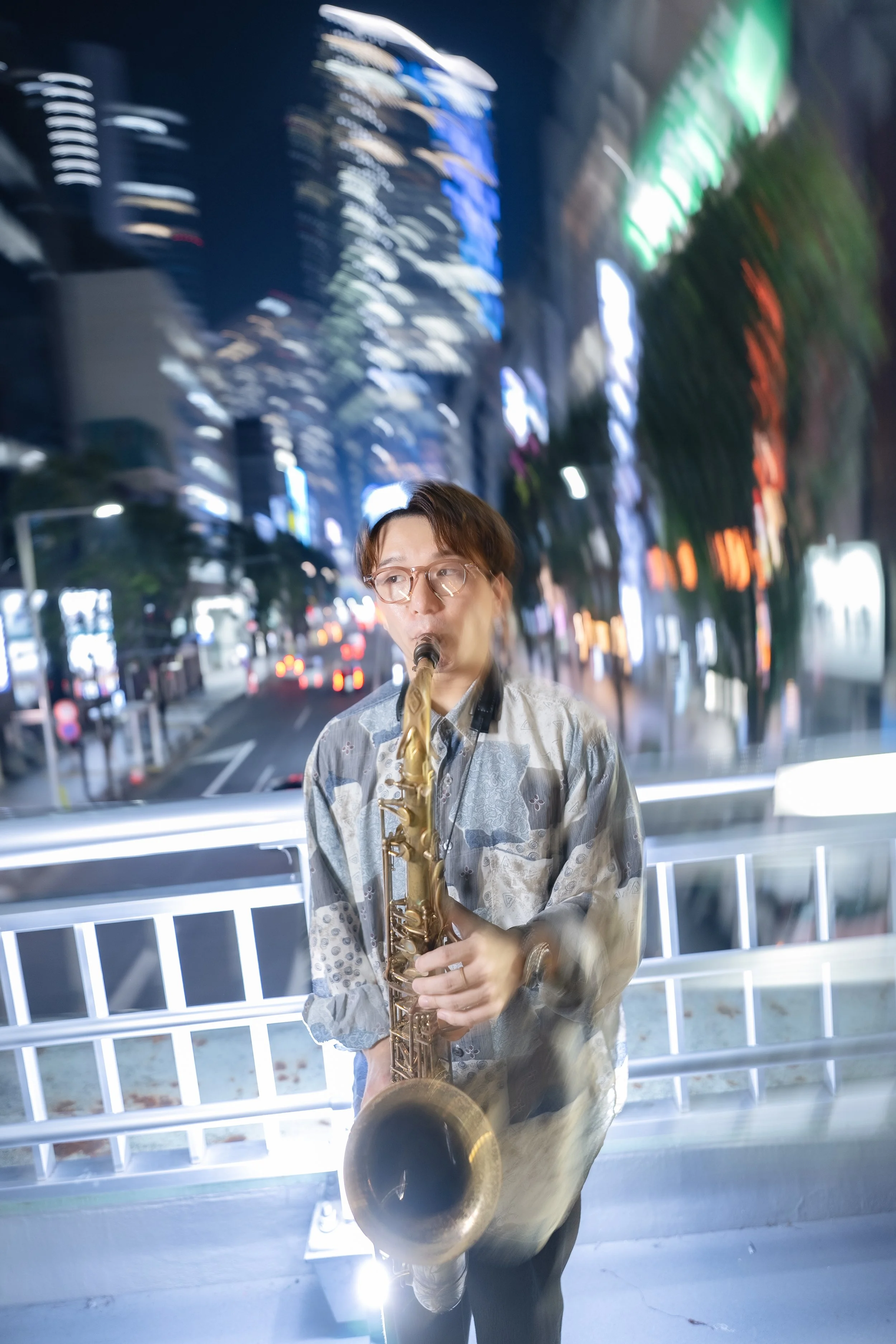 A person wearing glasses and a patterned shirt, playing a saxophone on a city street at night with illuminated buildings and signs in the background.