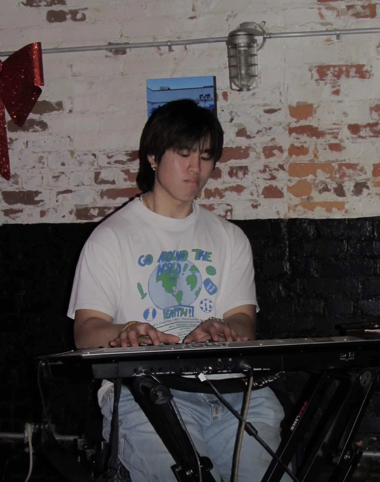 Young man playing a keyboard in a room with red brick and black painted wall, wearing a white T-shirt with a globe graphic and green text.
