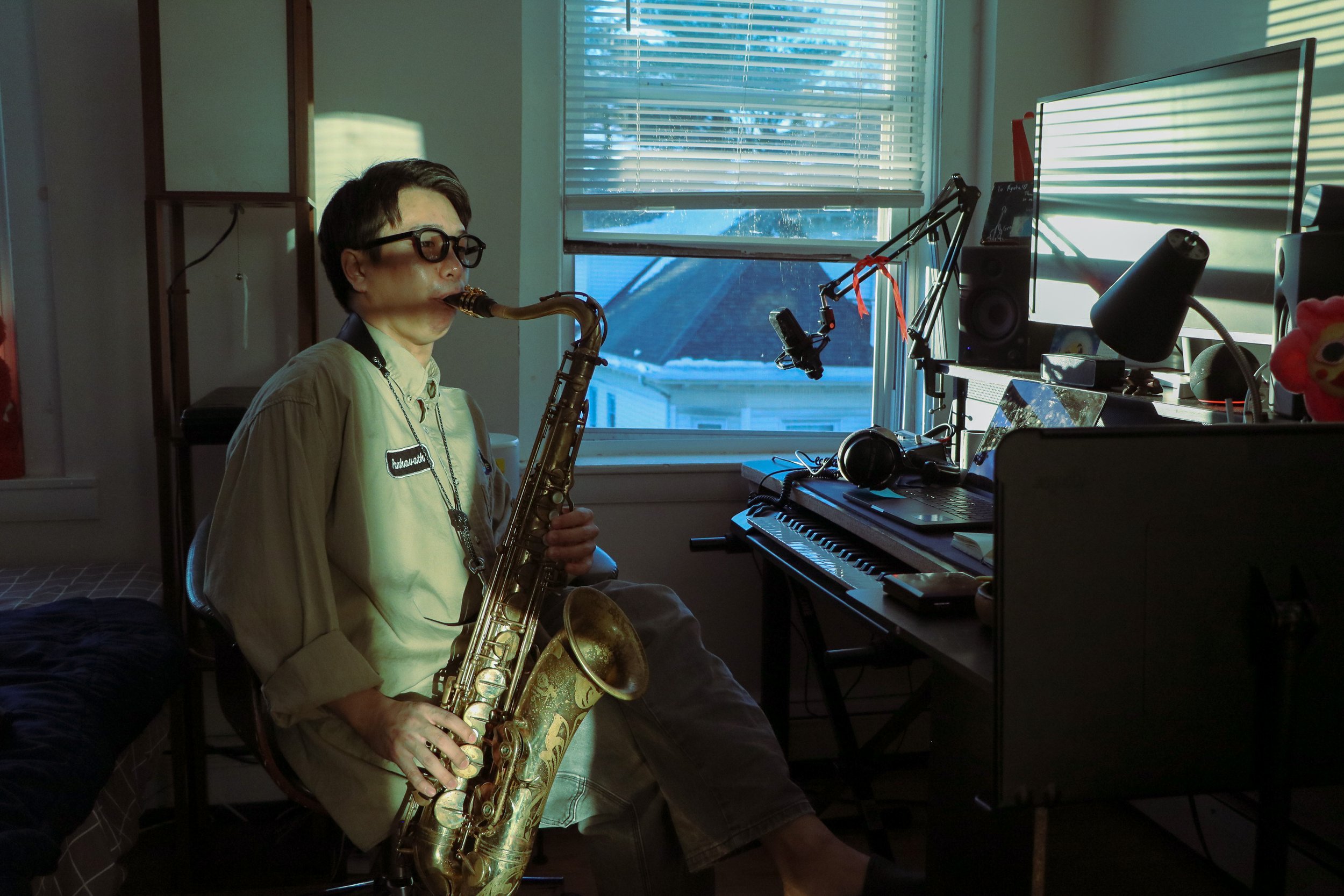 A person wearing glasses and a beige shirt playing a saxophone indoors near a window, with a computer monitor and speakers on a desk.