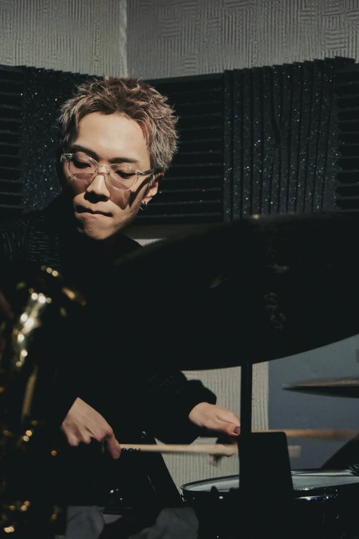 A person with light-colored hair, glasses, and earrings playing a drum set in a music studio