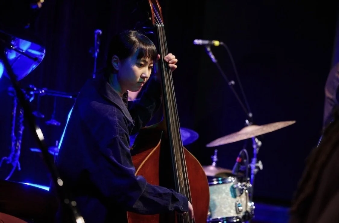 Young woman playing double bass on stage during a jazz performance, with drums and a microphone visible in the background.