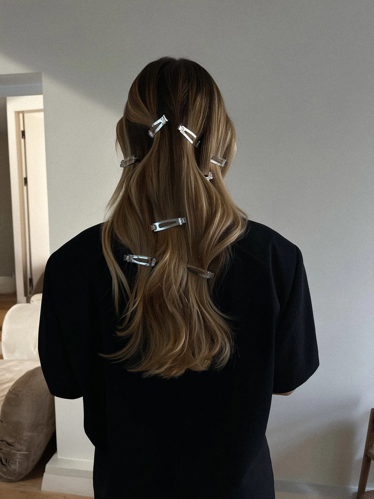 Back view of a woman with long, wavy, light brown hair secured with clear hair clips, wearing a black top, standing indoors against a plain gray wall.