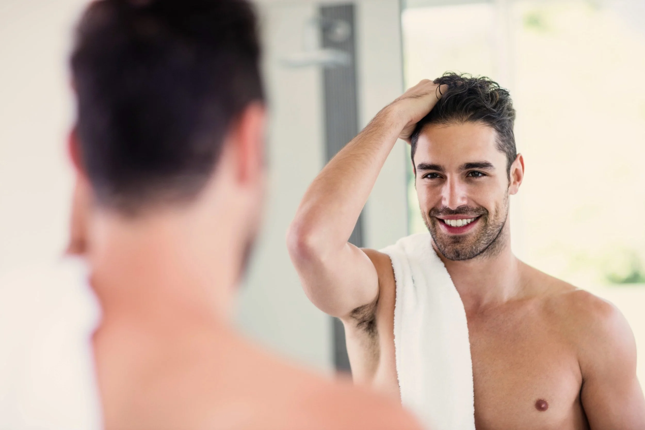 Young man smiling while looking at his reflection in the mirror, touching his hair.