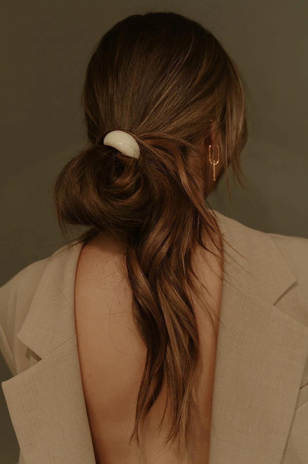 Back of a woman's head showing her brown hair styled in a low bun with a white hair clip, wearing a beige blazer and a gold earring, with her shoulder exposed.