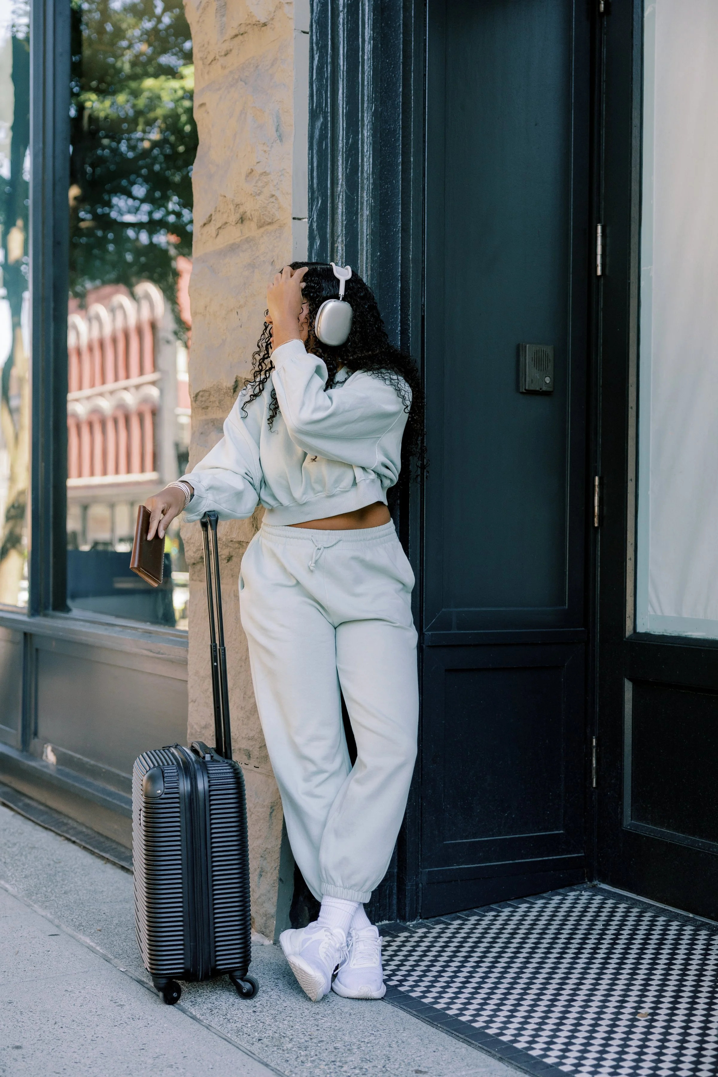 A woman wearing white sweatpants, white sneakers, and a white sweatshirt stands outside a building with a taxi window behind her. She has curly hair and is wearing large white headphones, holding a passport in one hand and a black suitcase with wheels on the ground beside her.