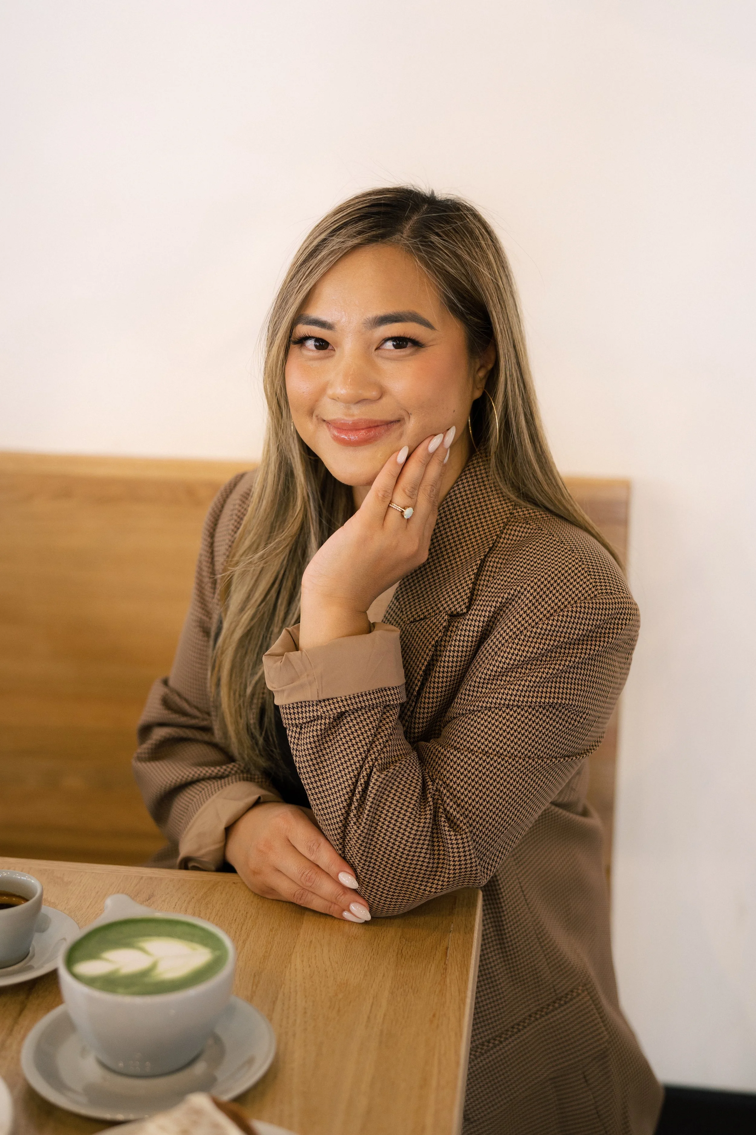 A woman with long hair, smiling, rests her chin on her hand with a ring on her finger, sitting at a wooden table with green matcha drinks in front of her, in an indoor setting with a light-colored wall.