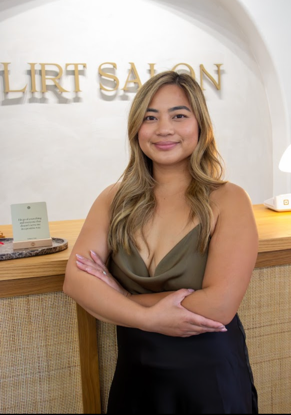 A young woman with long, wavy hair smiling and standing with her arms crossed in front of a hotel reception desk, with partial sign reading 'LIRT SALON' in the background.
