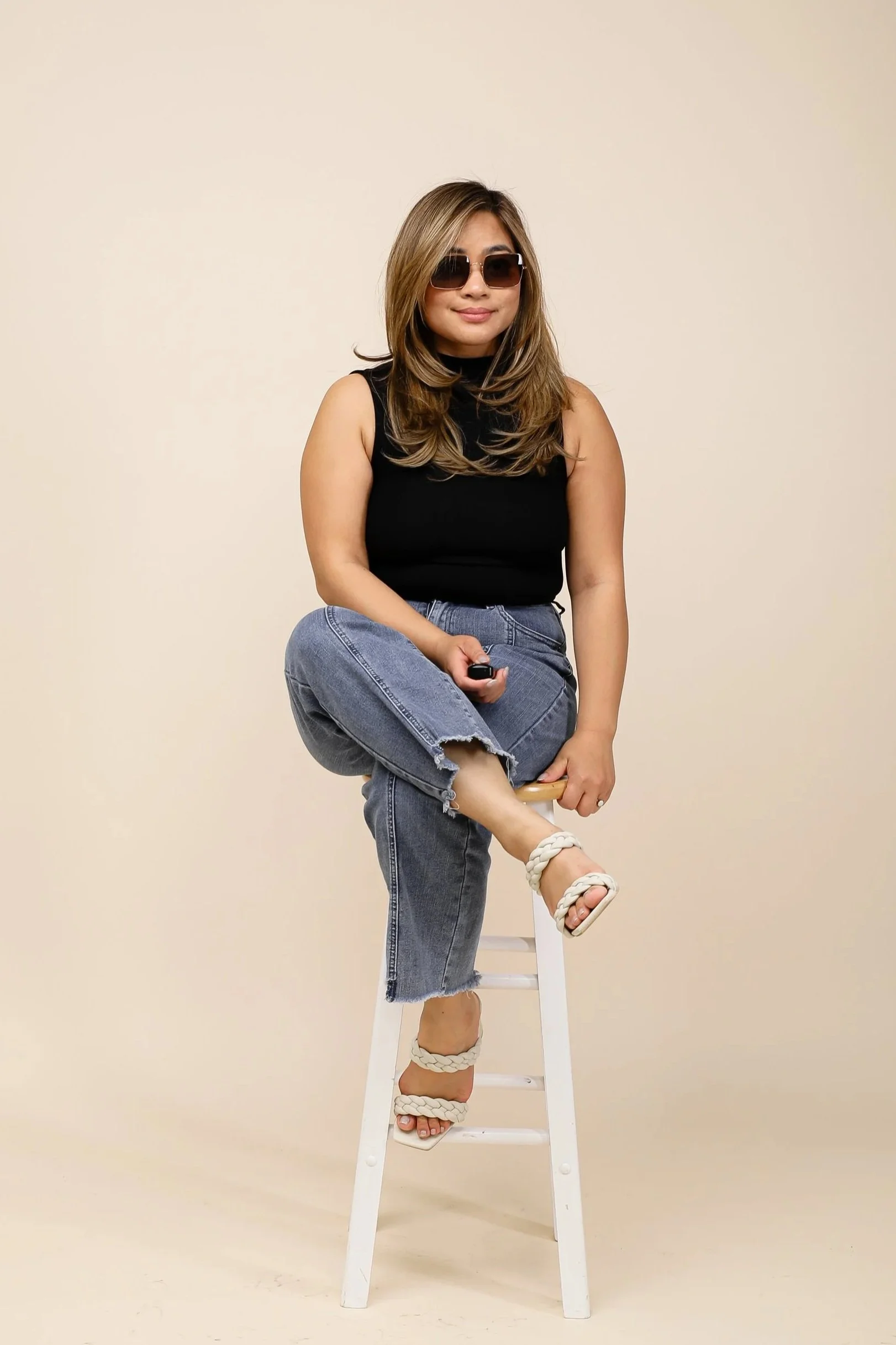 A woman with long brown hair wearing dark sunglasses, a black sleeveless top, and ripped blue jeans. She is sitting on a white stool, holding a remote control, with one leg crossed over the other, wearing sandals with braided straps, in front of a beige background.