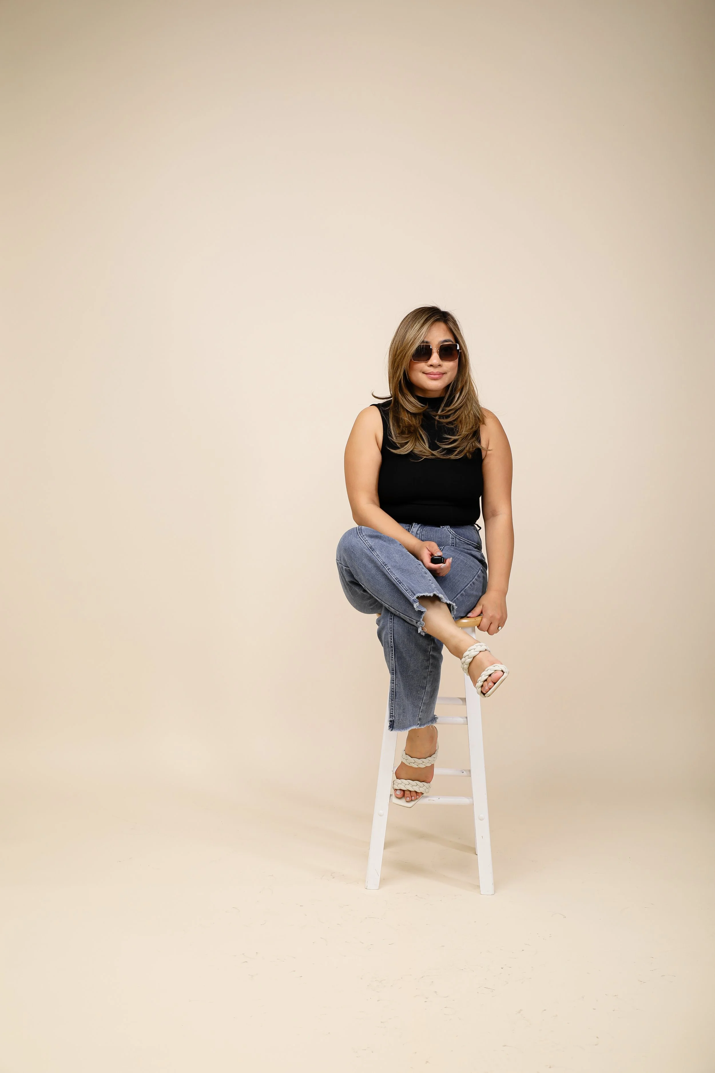 A woman with long hair, wearing sunglasses, a black sleeveless top, and ripped jeans, is sitting on a white stool with one foot resting on a rung, holding a black remote, against a plain beige background.