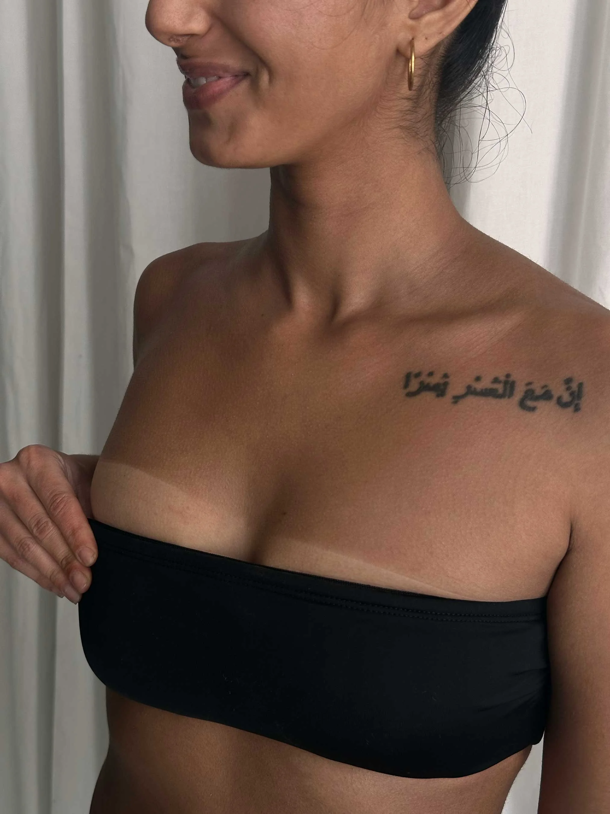 Close-up of a woman with a tattoo on her shoulder, wearing a black top, holding her shoulder with her hand to show the difference her sunless tan made.