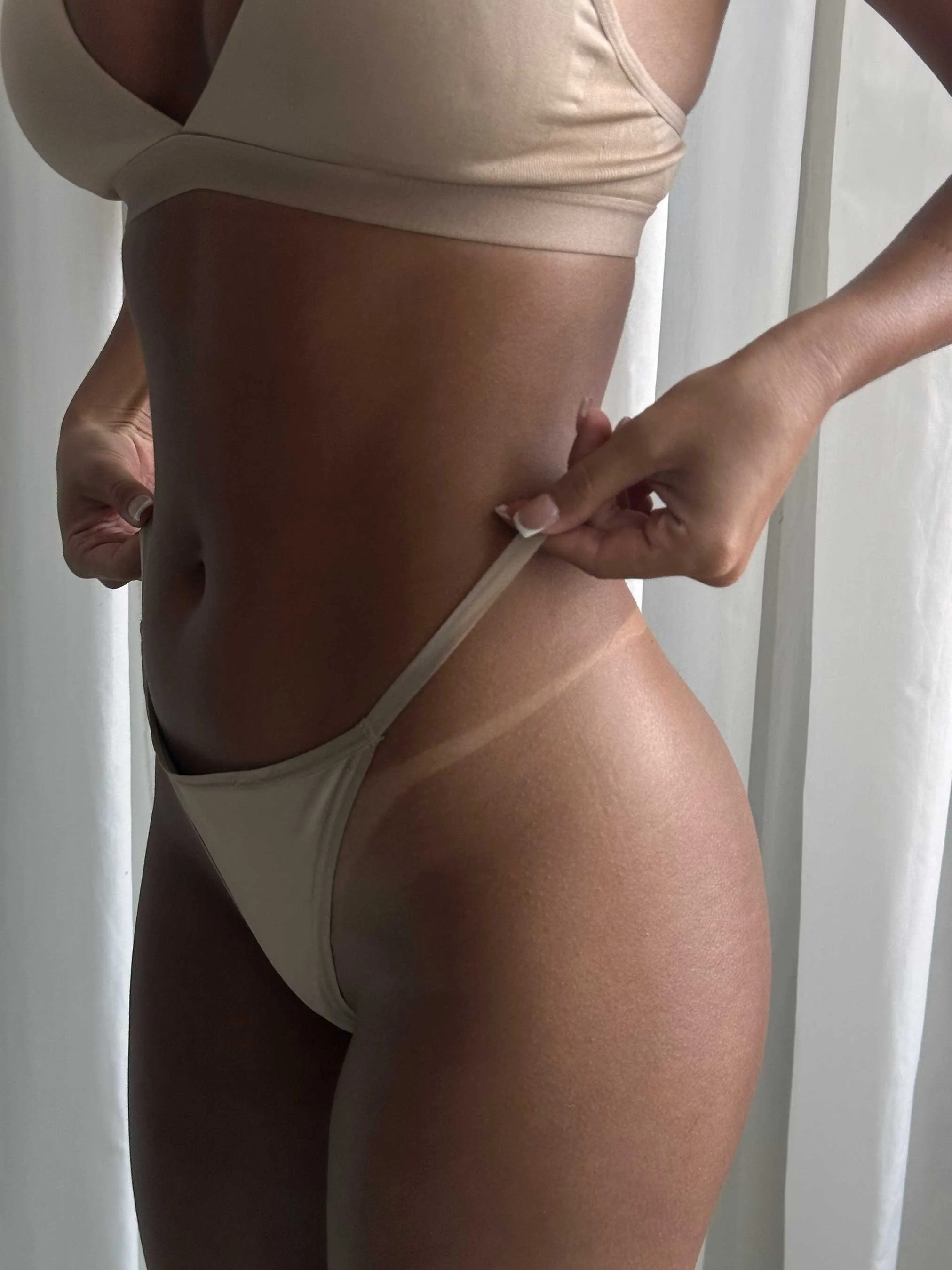 Close-up of a woman's midsection, showing her wearing a beige crop top and beige underwear revealing here sunless tan results.