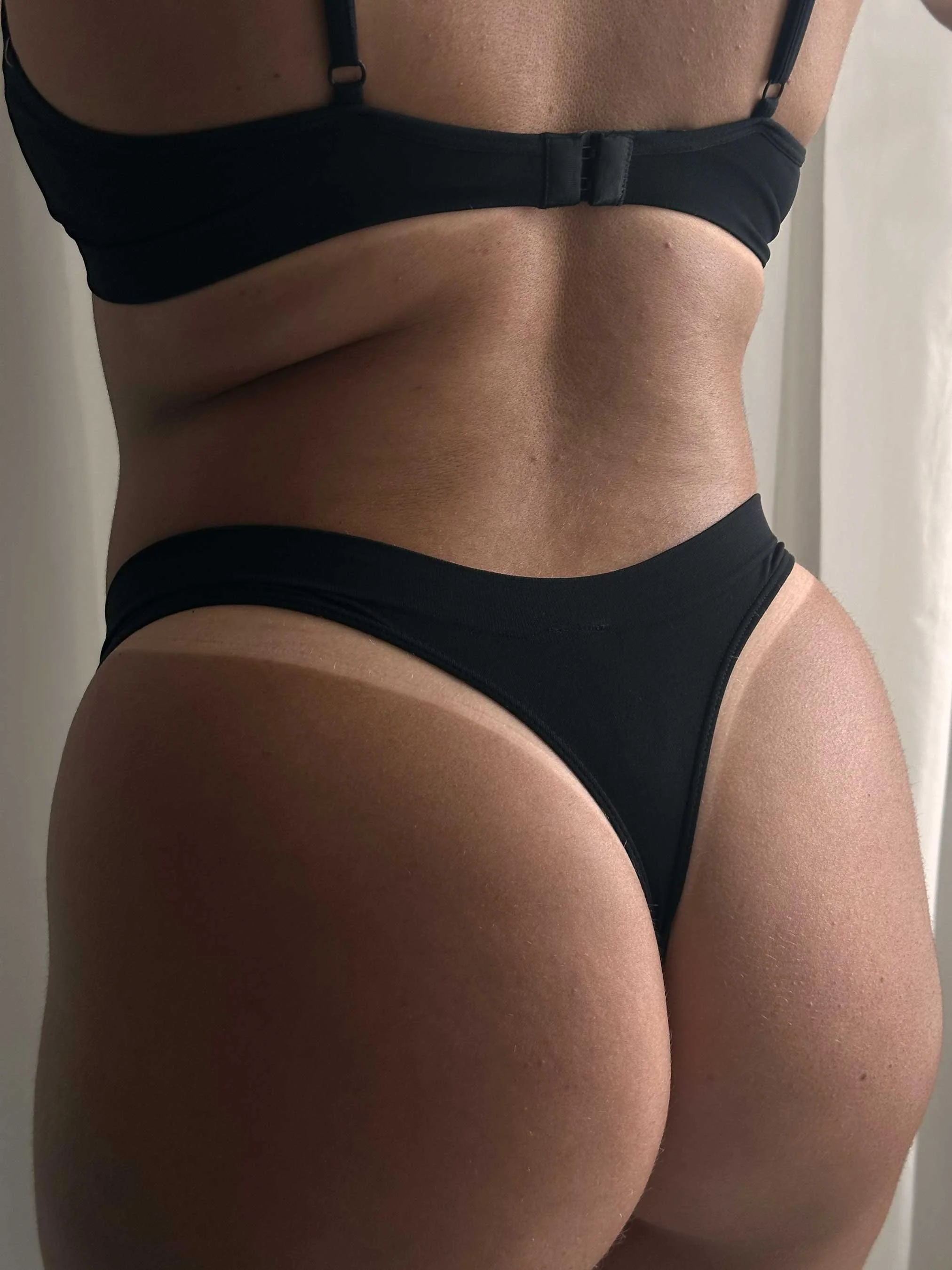 Close-up of a woman's lower back and hips, wearing black underwear and a black bra, revealing spray tan results.