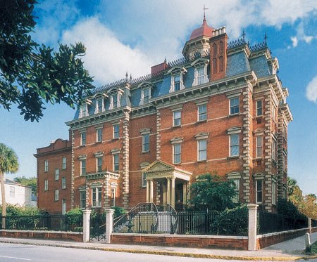 A large, historic brick mansion with ornate architecture, surrounded by a fence and trees, under a partly cloudy sky.
