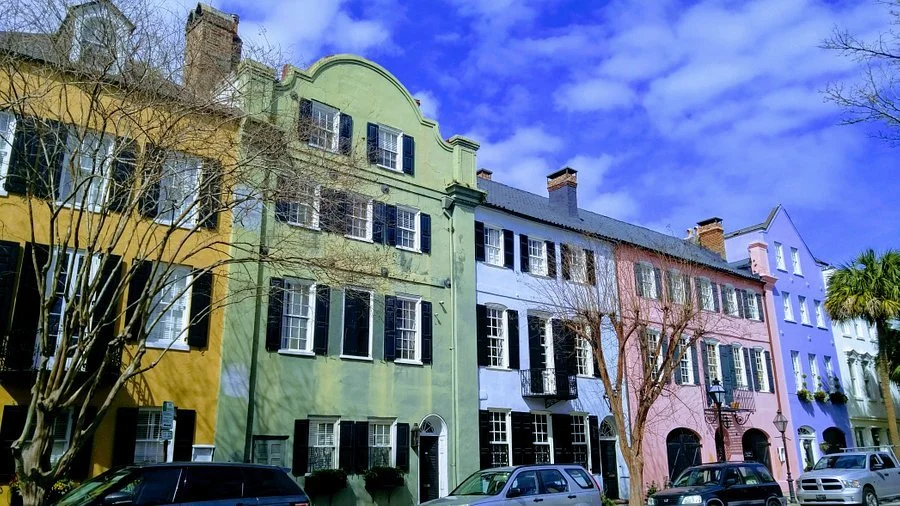 Colorful row of townhouses in pastel shades of yellow, green, blue, pink, and purple with cars parked in front and a partly cloudy blue sky above.