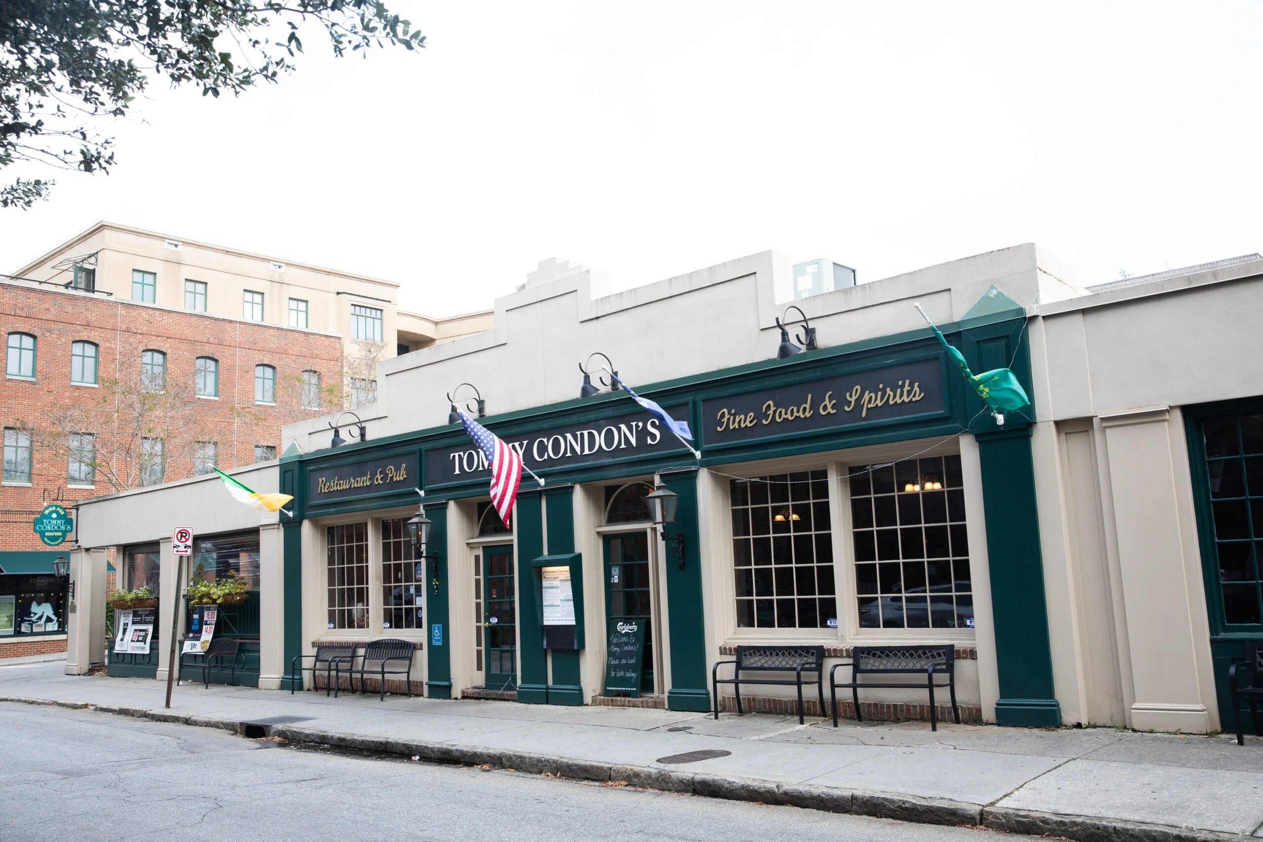 Exterior of Tony Conson's restaurant and pub with flags, benches outside, and a black sign with gold lettering. The building has large windows and a green awning with gold writing that says 'Fine Food & Spirits' and another sign that reads 'Restaurant & Pub'.