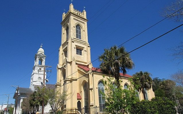 A historic church with a tall beige tower, arched windows, and a red roof, surrounded by palm trees and greenery, under a clear blue sky.