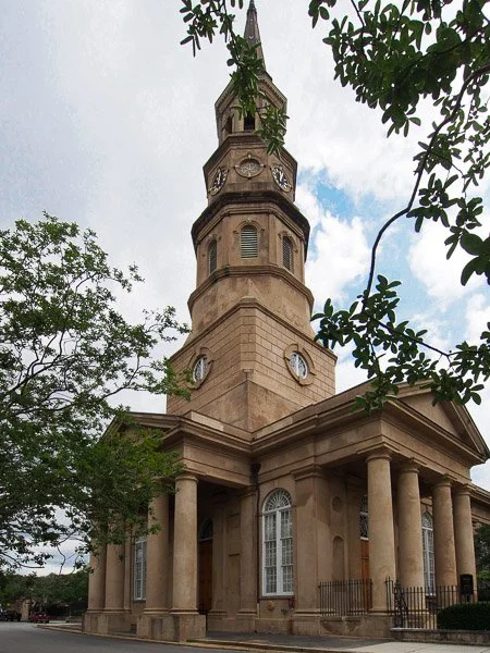 A historic church with a tall clock tower, columns at the entrance, and arched windows, surrounded by trees on a cloudy day.