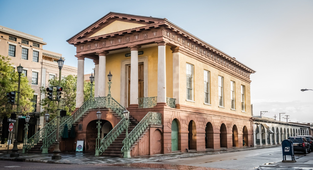 Historic yellow building with pink accents, columns, and stairs, located on a city street with other buildings and trees.