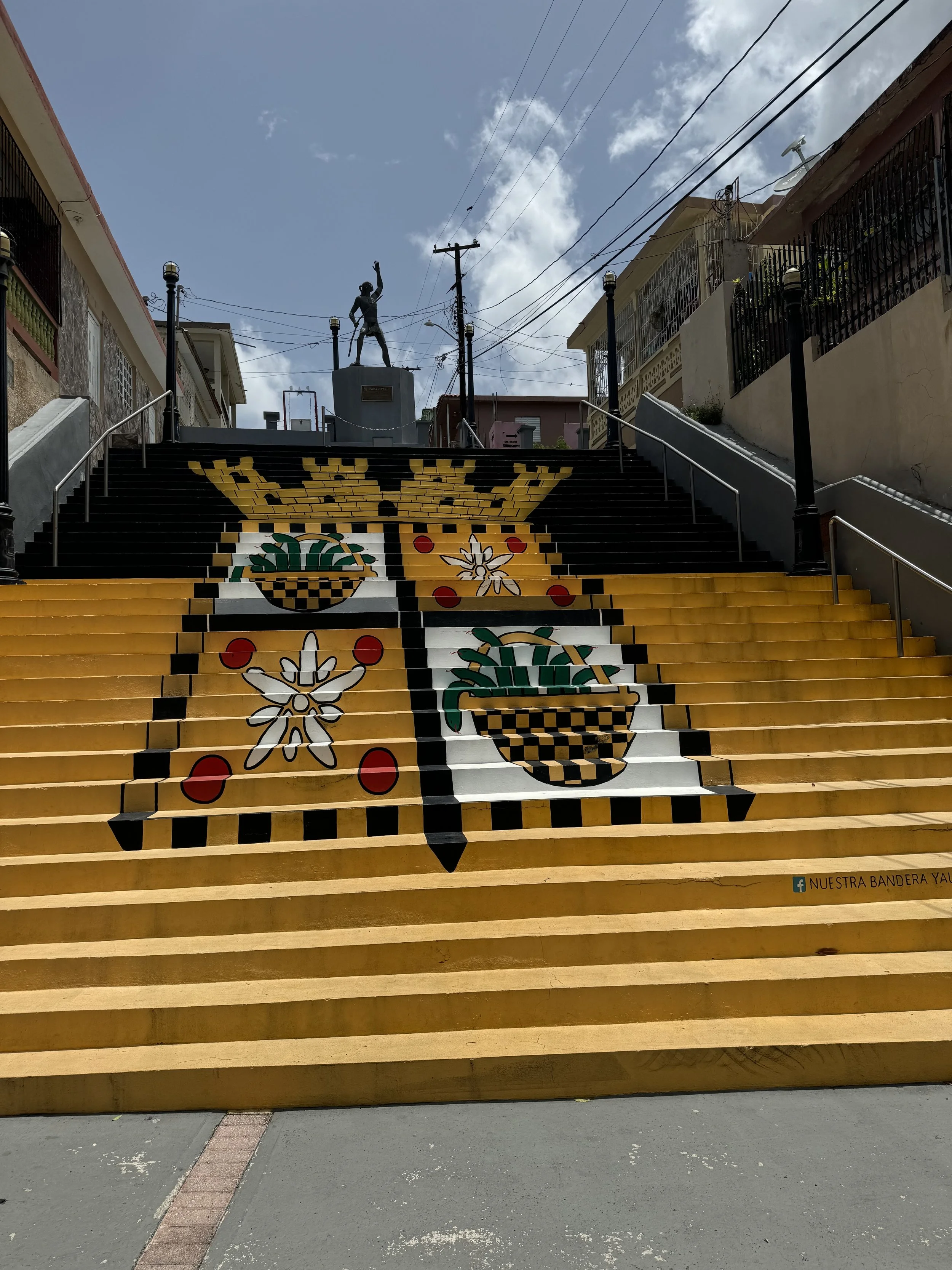 Colorful mural painted on the steps of an outdoor staircase depicting traditional Mexican symbols, including potted plants, flowers, and geometric patterns, with buildings and utility poles in the background.