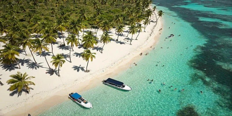 Aerial view of a tropical beach with palm trees, clear turquoise water, and boats with people swimming and relaxing in the water.