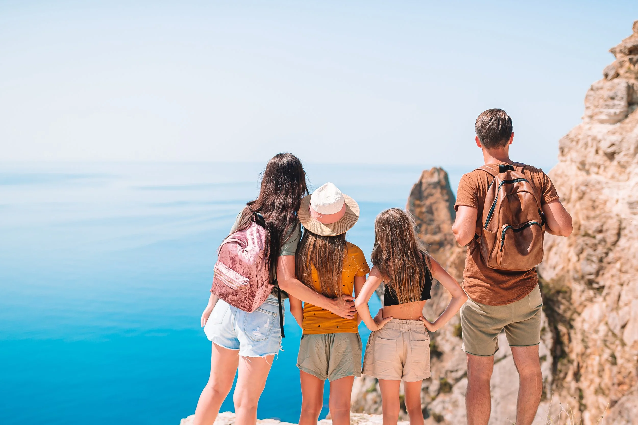 A family of five stands on a cliff overlooking the ocean, with rocky cliffs in the background, on a sunny day.