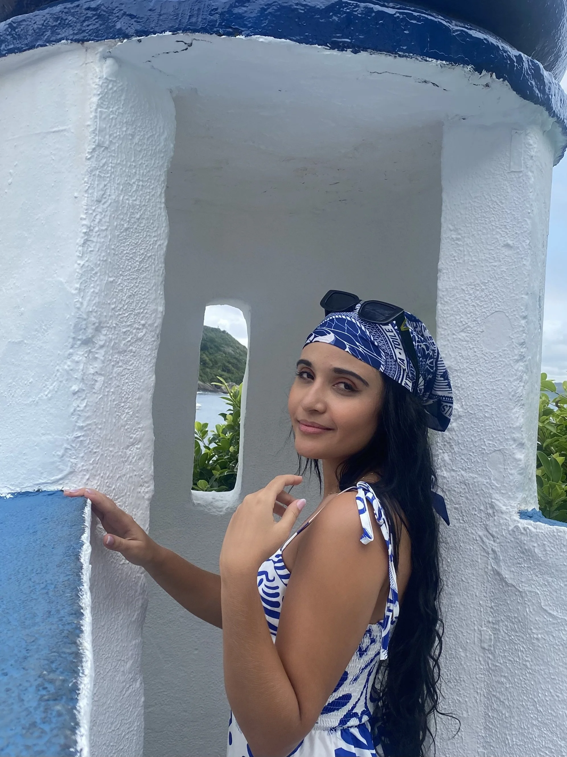 A young woman with long dark hair wearing a blue and white patterned dress and matching headscarf, standing inside a small white structure with blue accents, outdoors with green foliage and water in the background.