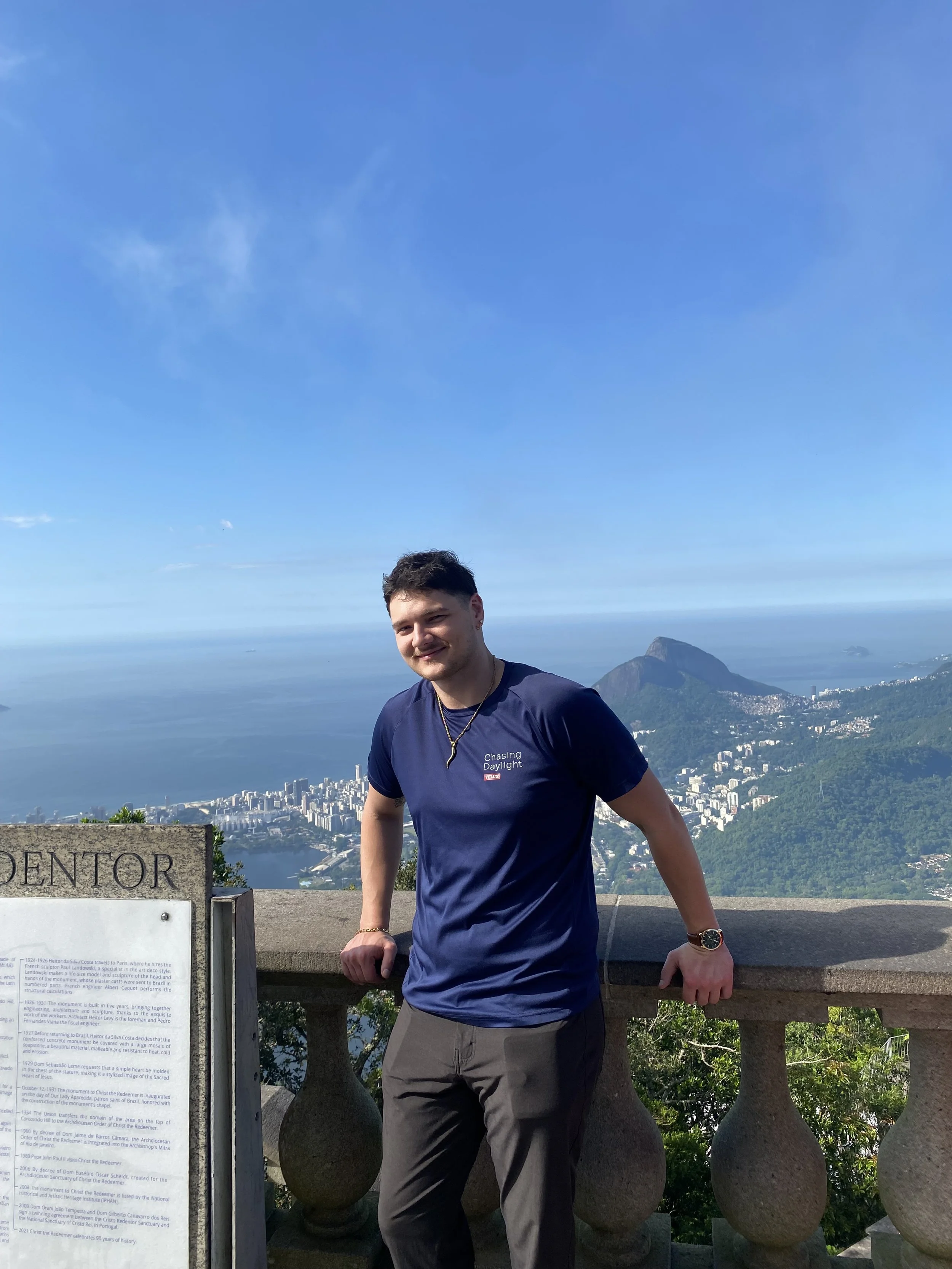 Young man standing outdoors on a scenic viewpoint with a panoramic view of the city, mountains, and ocean in the background, on a clear day.
