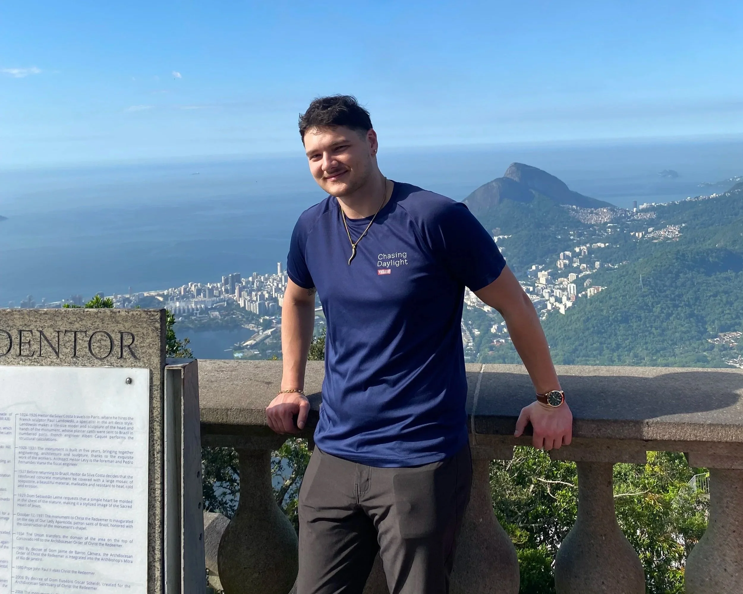 A young man in a navy blue T-shirt and gray shorts standing at a scenic overlook with a view of Rio de Janeiro, Brazil, including Sugarloaf Mountain and the city skyline, on a clear day.
