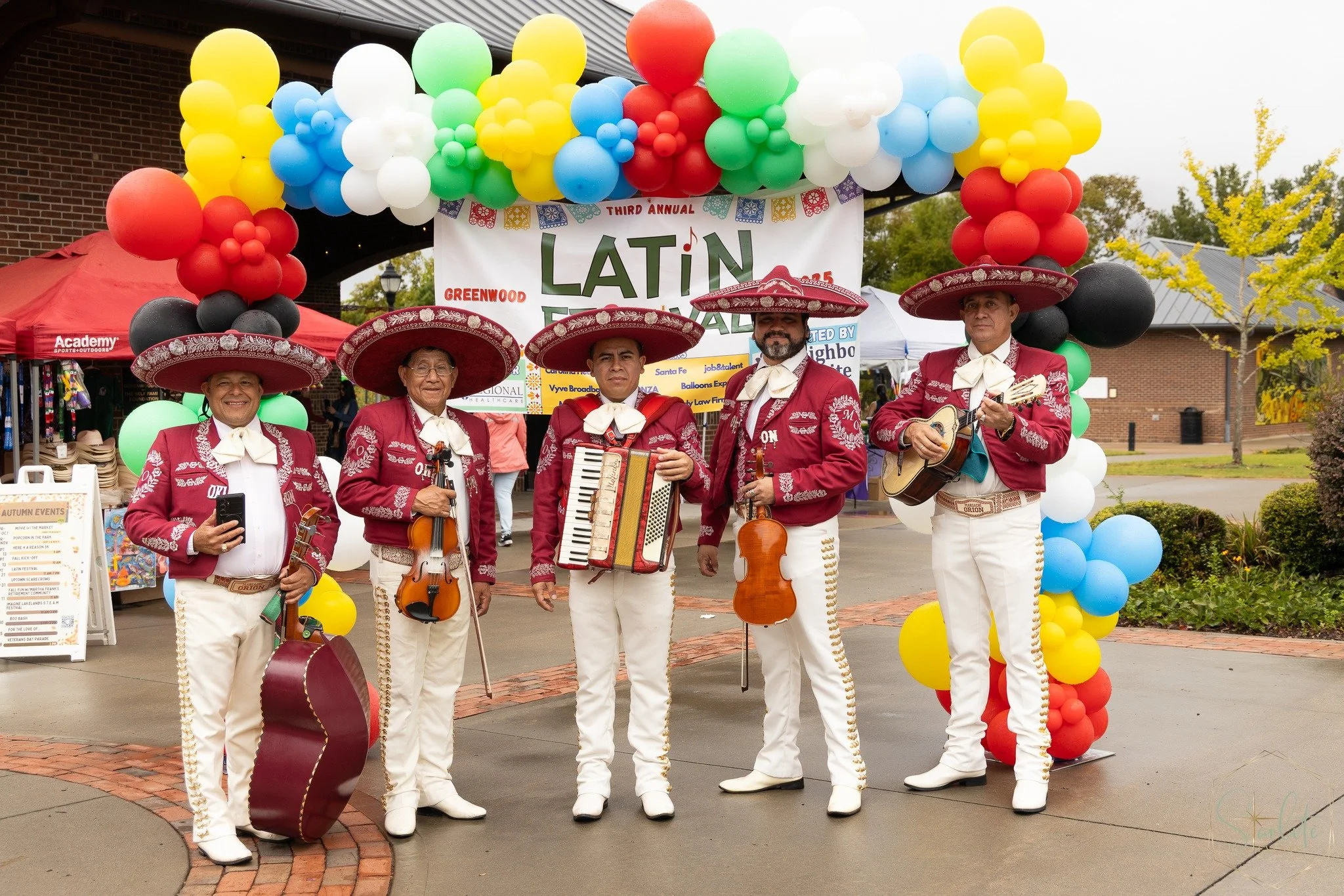 Five Mariachi musicians in traditional red and white outfits with large sombreros, holding instruments, standing in front of a colorful balloon arch and a banner at a Latin festival event.