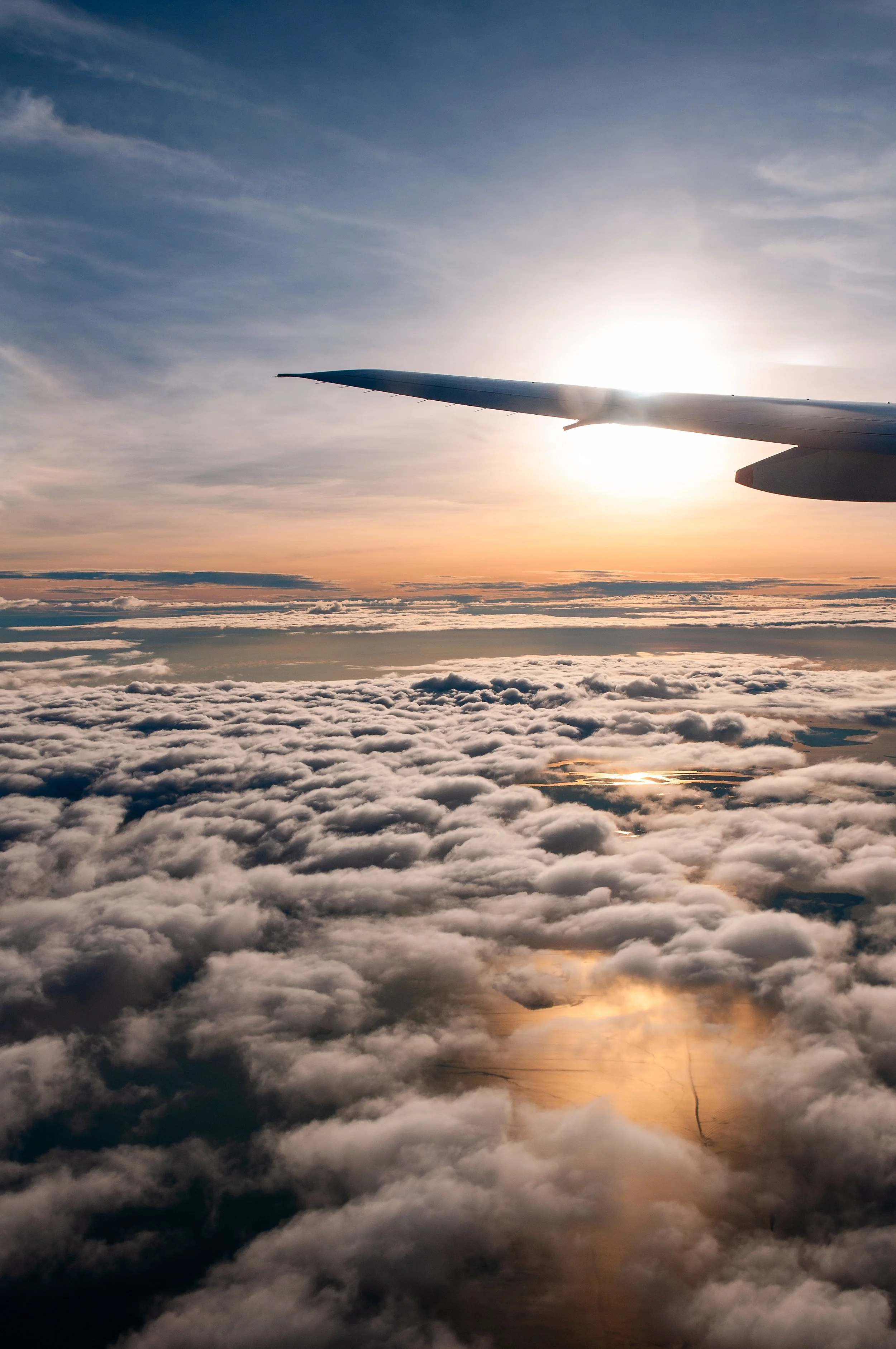 View of an airplane wing with the sun setting or rising, above a sky full of clouds and reflections on water below.