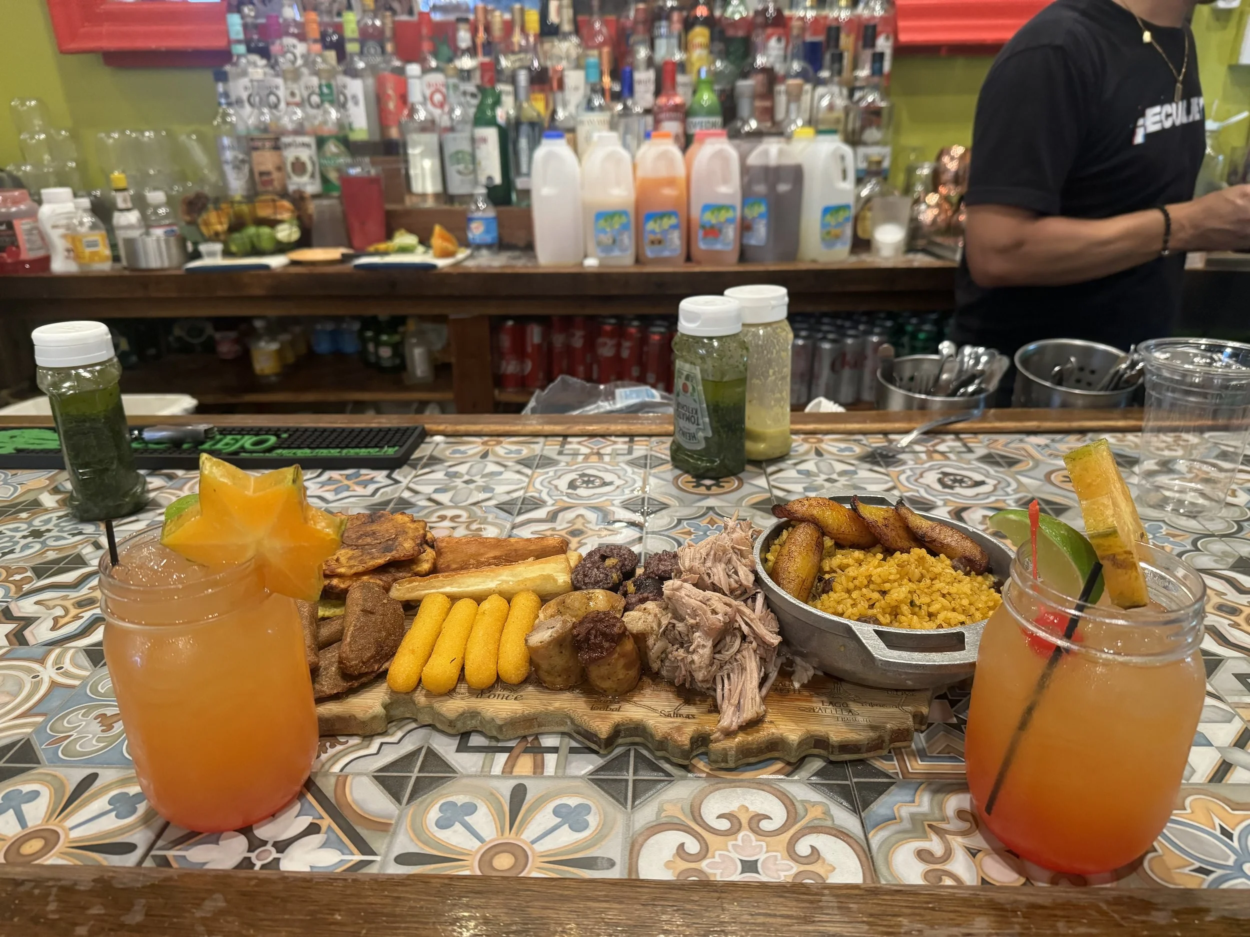A colorful Mexican-style meal with two margaritas, grilled meats, rice, fried plantains, and sides served on a decorative tile table in a restaurant setting.