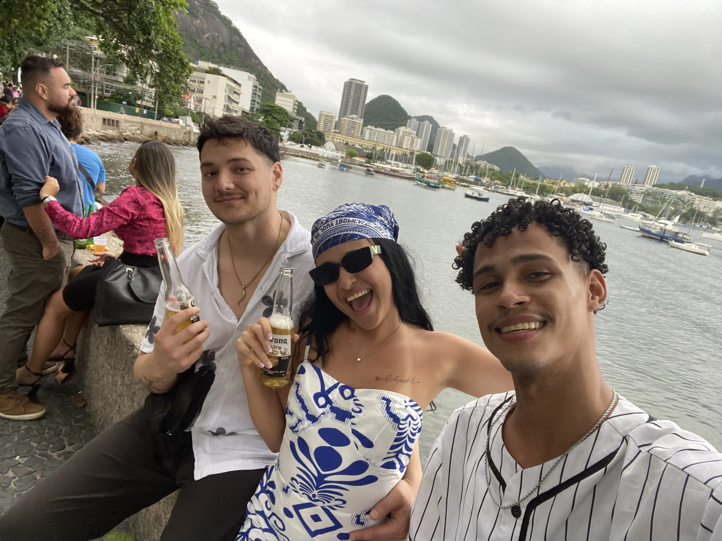Three friends taking a selfie near a waterfront with boats and a cityscape in the background. They are holding drinks and smiling at the camera, with cloudy skies overhead.