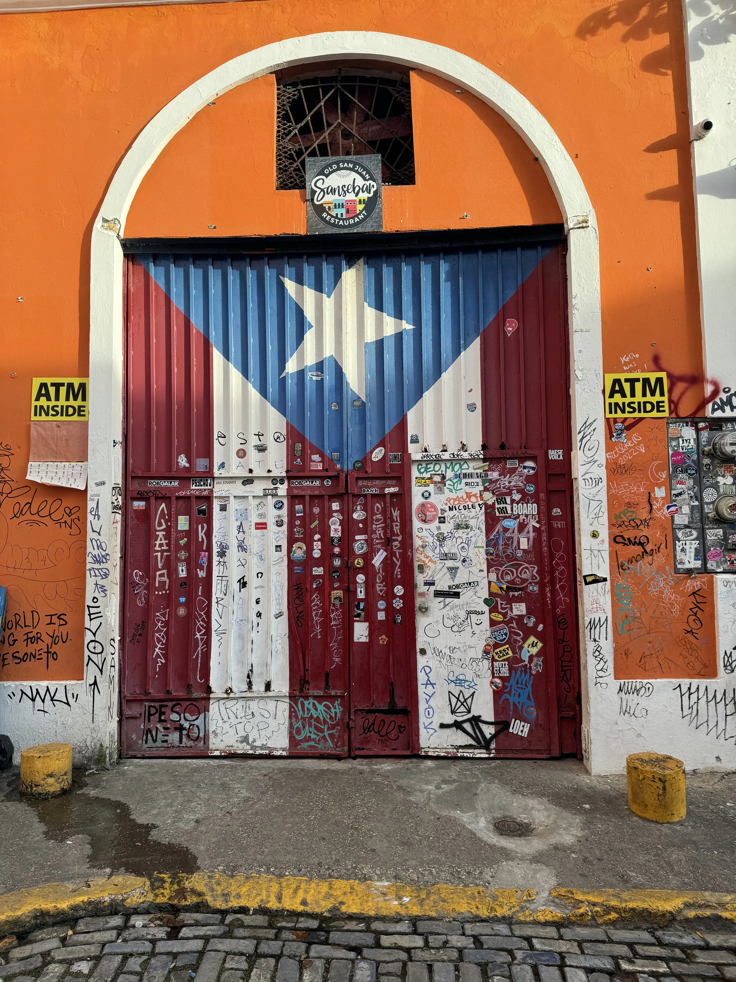 A gate painted with the Puerto Rican flag, featuring graffiti and stickers, flanked by orange walls with yellow 'ATM INSIDE' signs on each side.