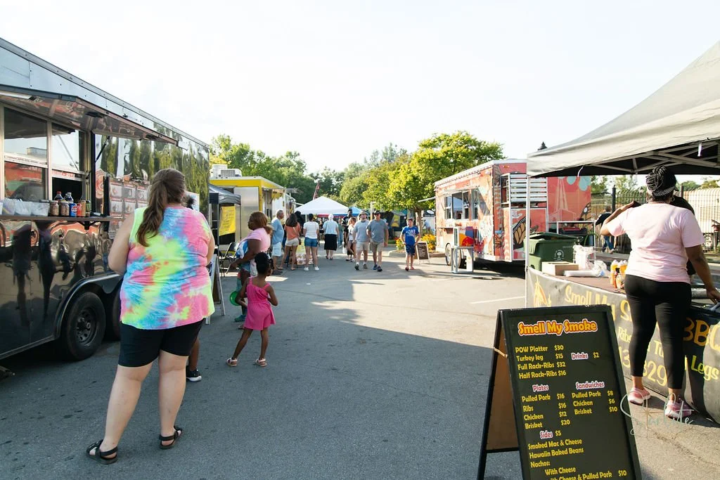People browsing food trucks and stands at an outdoor event field on a sunny day.