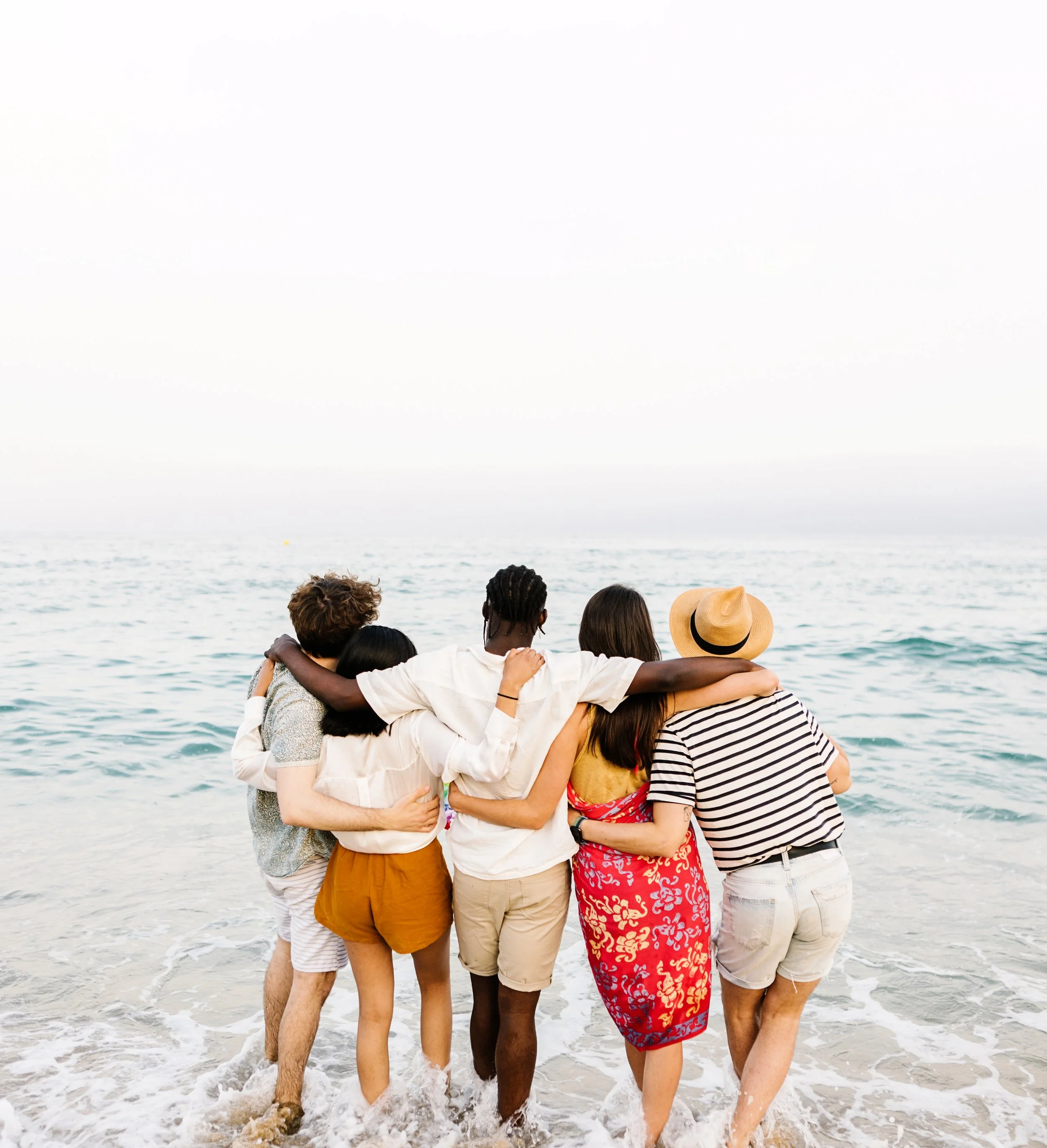 A group of five friends standing on the beach, facing the ocean with arms around each other.