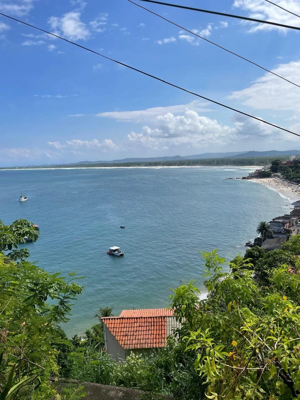 Scenic view of a coastal area with a blue sky, scattered clouds, a calm sea with boats, a sandy beach, and some houses with red-tiled roofs among green foliage.
