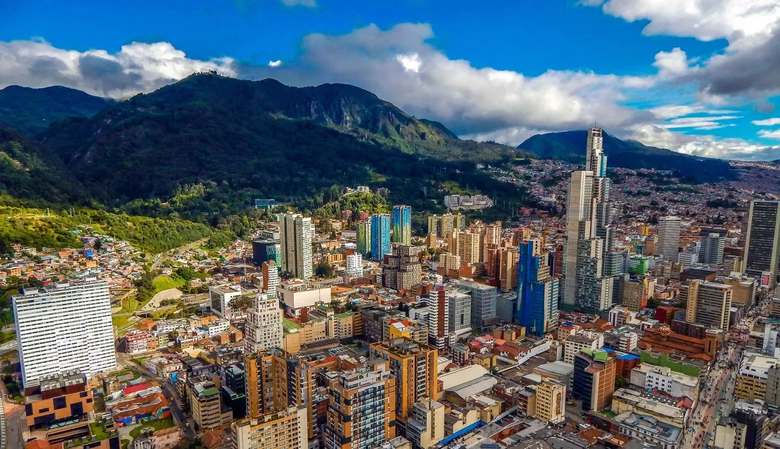 A cityscape of a bustling urban area with numerous high-rise buildings, surrounded by green hills and mountains in the background, with a cloudy sky overhead.