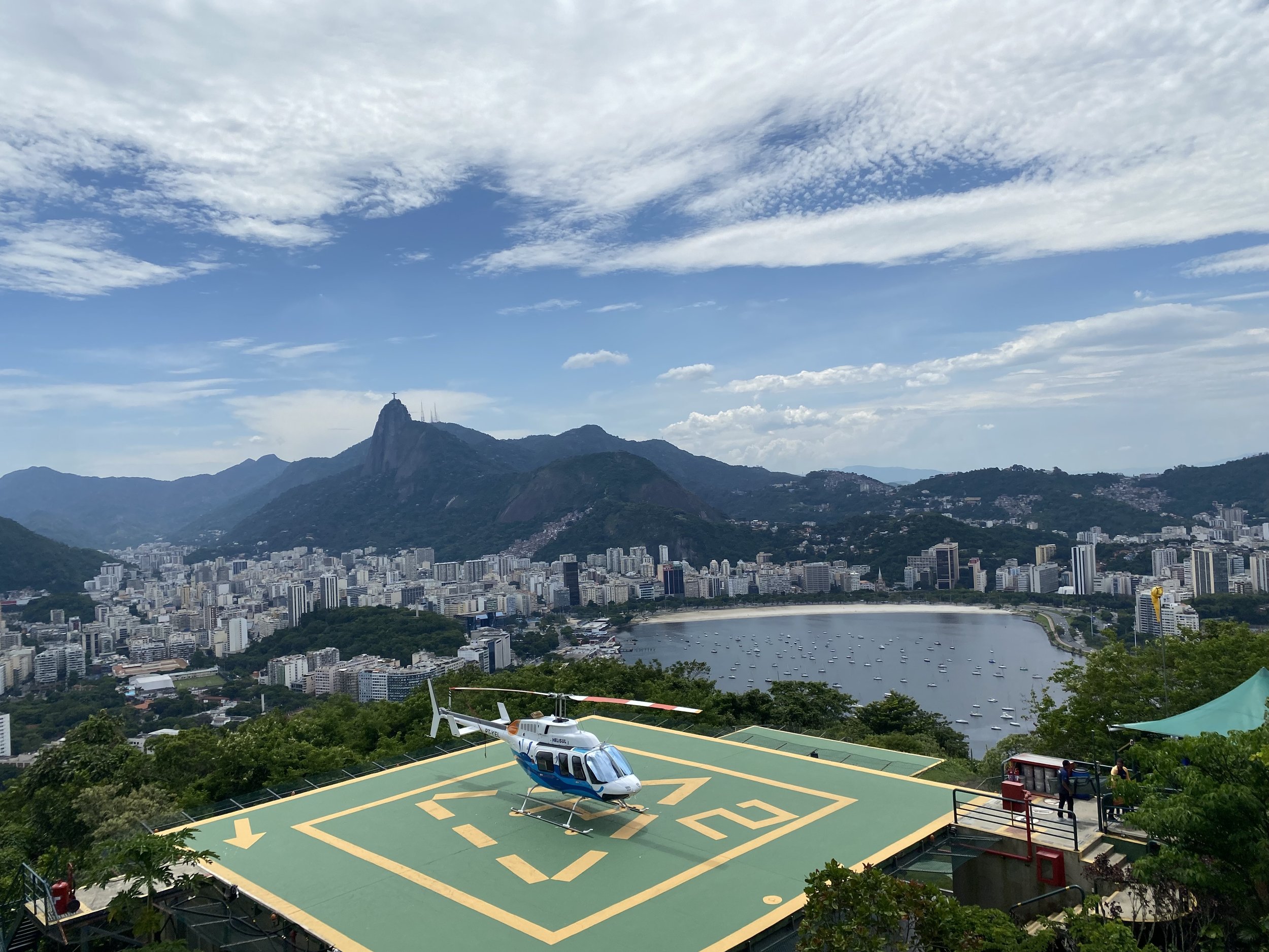 Helipad with a blue and white helicopter on a hilltop overlooking Rio de Janeiro, Brazil, featuring mountains, city buildings, and a bay with boats.