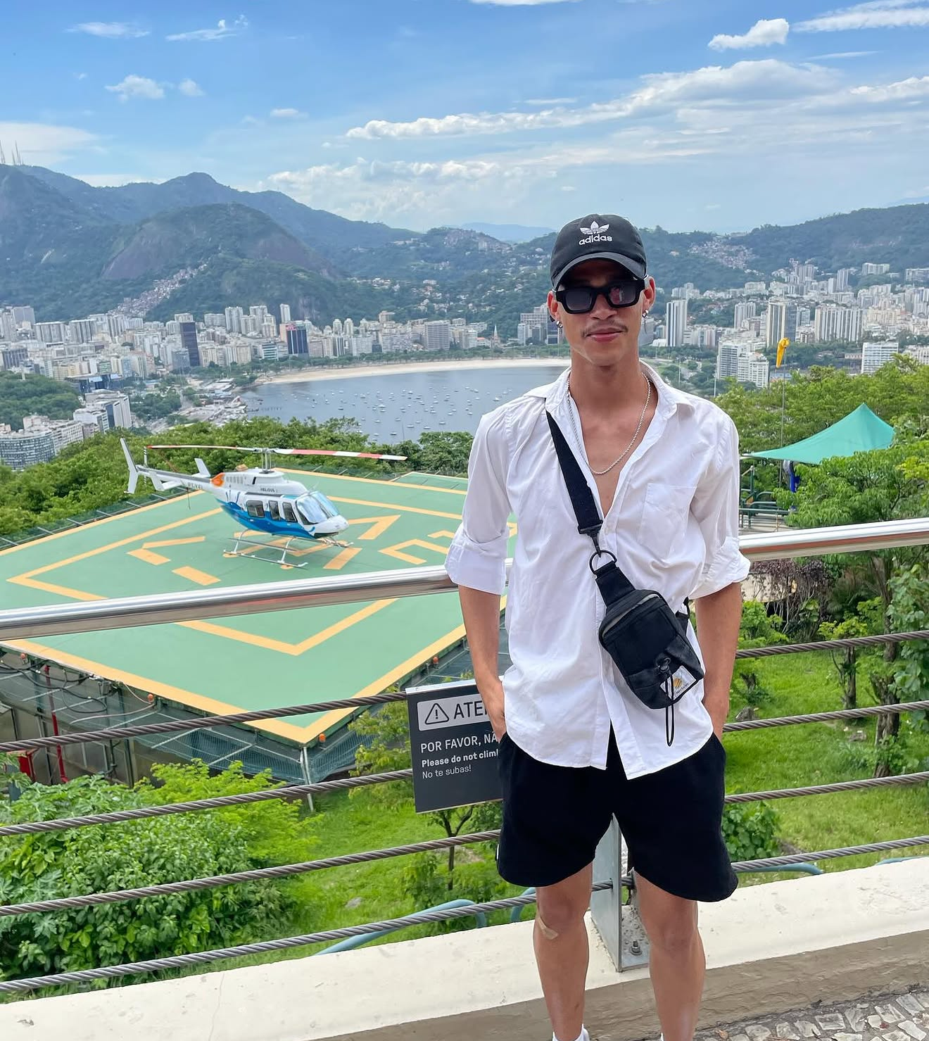 Young man in sunglasses, black cap, white shirt, and black shorts standing outdoors with a cityscape, mountains, and a helipad with a helicopter in the background.